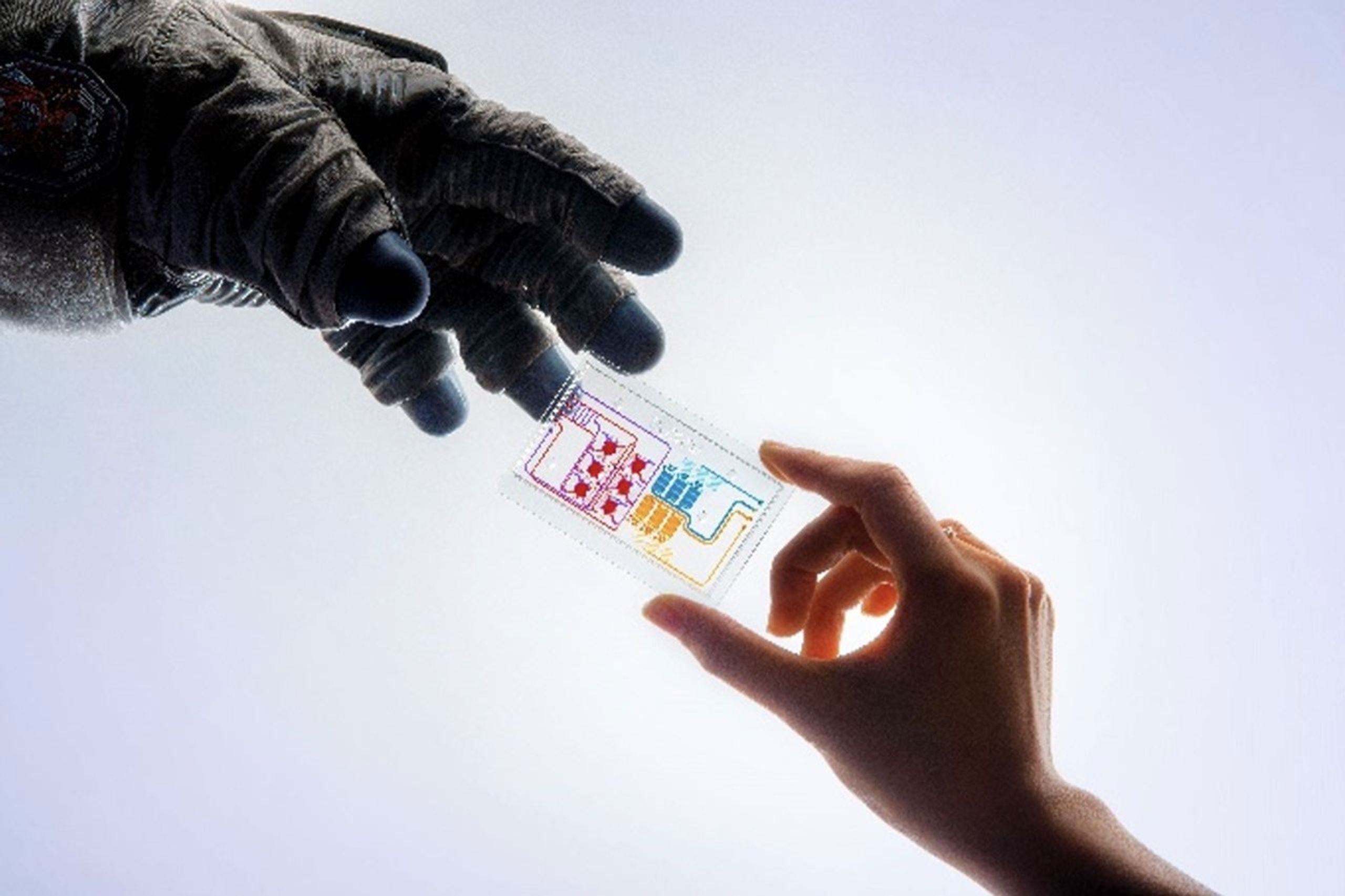 Un fondo blanco brillante proporciona un escenario claro para esta imagen fotografiada en estudio. La mano enguantada de un astronauta se extiende desde la izquierda hacia una mano que sostiene un chip de tejido a la derecha. El chip de tejido es un dispositivo transparente del tama�o de una tarjeta de cr�dito que contiene redes de puertos y canales rojos, azules y amarillos. Estos peque�os dispositivos podr�an ayudarnos a dar el siguiente gran salto y beneficiar la salud humana en la Tierra. Cr�dito: Unive