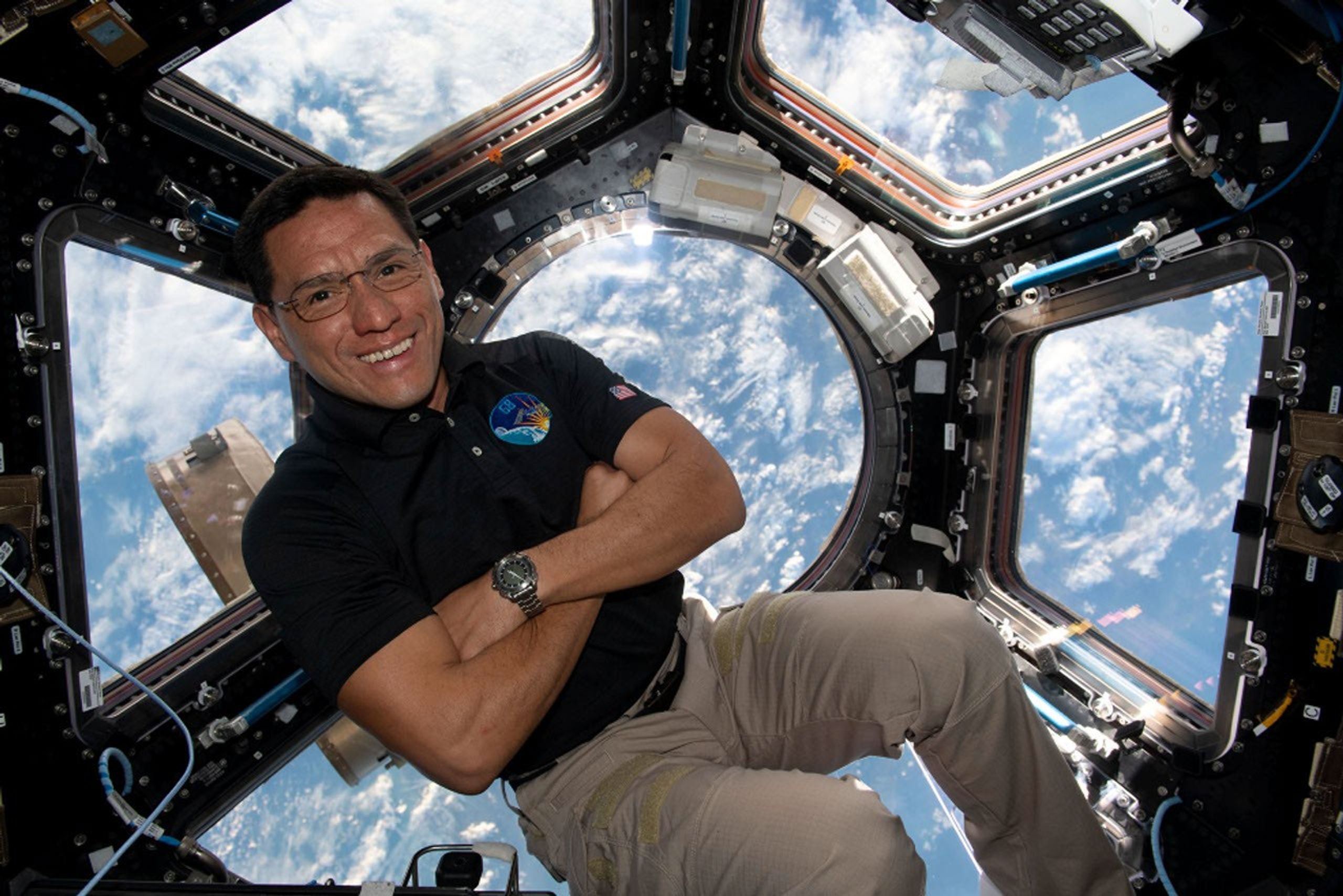 Imagen de Frank Rubio flotando en la c�pula de observaci�n de la Estaci�n Espacial Internacional. Rubio, un hombre de pelo y ojos oscuros y con lentes, sonr�e y tiene los brazos cruzados. Va vestido con un polo oscuro y pantalones khaki. Detr�s suyo se ve la atm�sfera de la Tierra a trav�s de las ventanas de observaci�n de la c�pula.