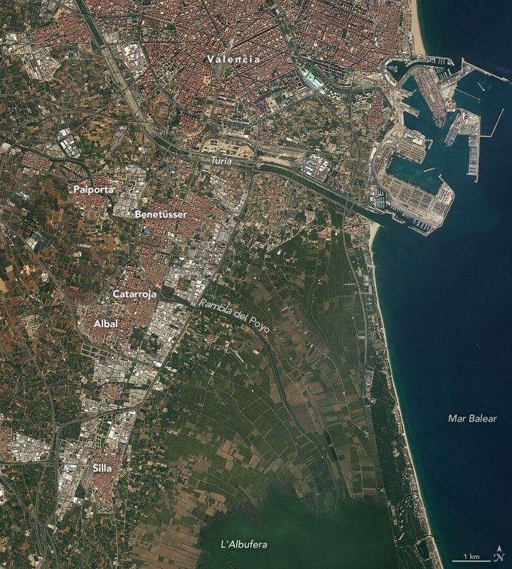 Esta imagen satelital de octubre de 2022 muestra Valencia, España, antes de las lluvias torrenciales de octubre de 2024. La imagen muestra varias ciudades con grupos de edificios rojos y blancos a la izquierda de la imagen y tierras agrícolas verdes a la derecha. El mar Balear está a la derecha de la imagen y se ve un puerto en la esquina superior izquierda.