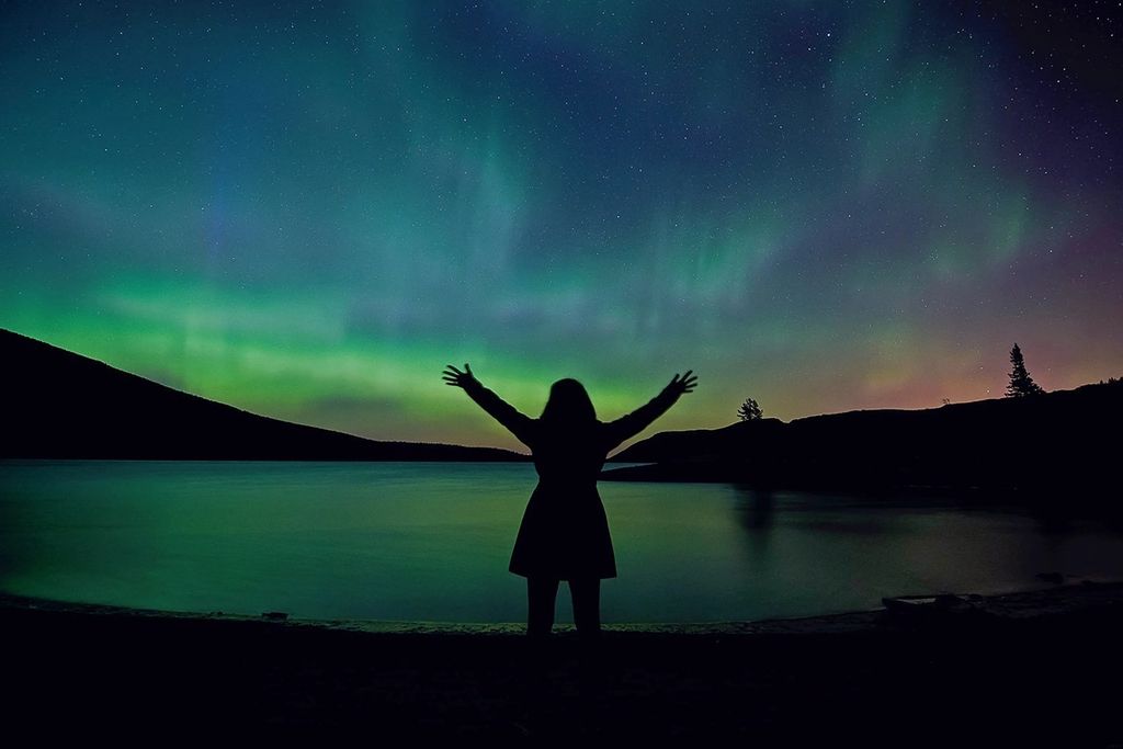 Fotografía de la figura de una mujer de pie con los brazos en alto hacia una colorida aurora que se muestra frente a ella.  Una masa de agua refleja la aurora en la parte inferior de la imagen; al fondo se ven algunas colinas. Es de noche. 