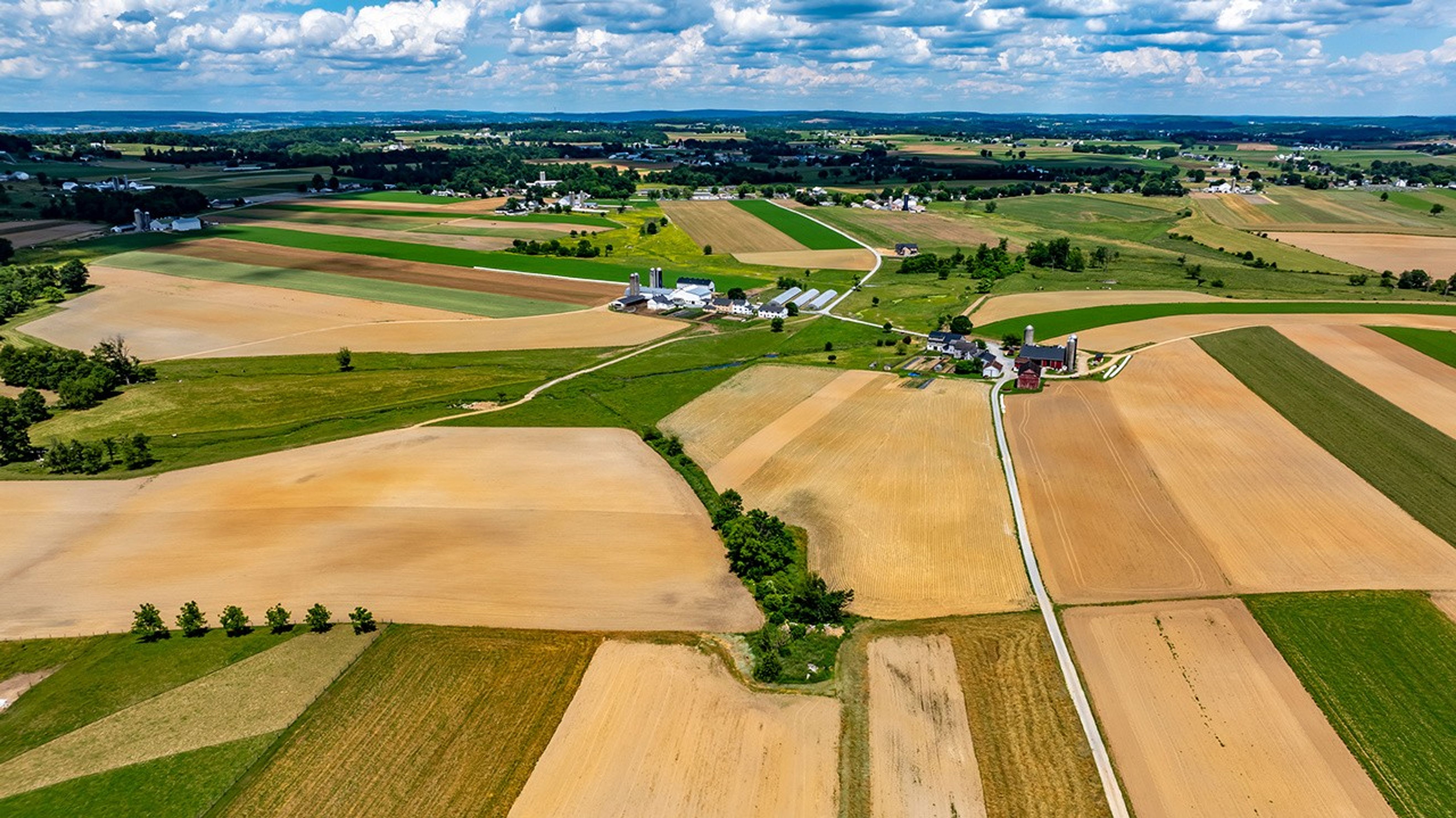 Por el paisaje se extienden frondosos campos de diversos tonos de verde y marrón, salpicados de granjas. La amplia vista revela una zona rural con una carretera despejada que atraviesa las tierras de cultivo. El cielo celeste está salpicado de nubes blancas.