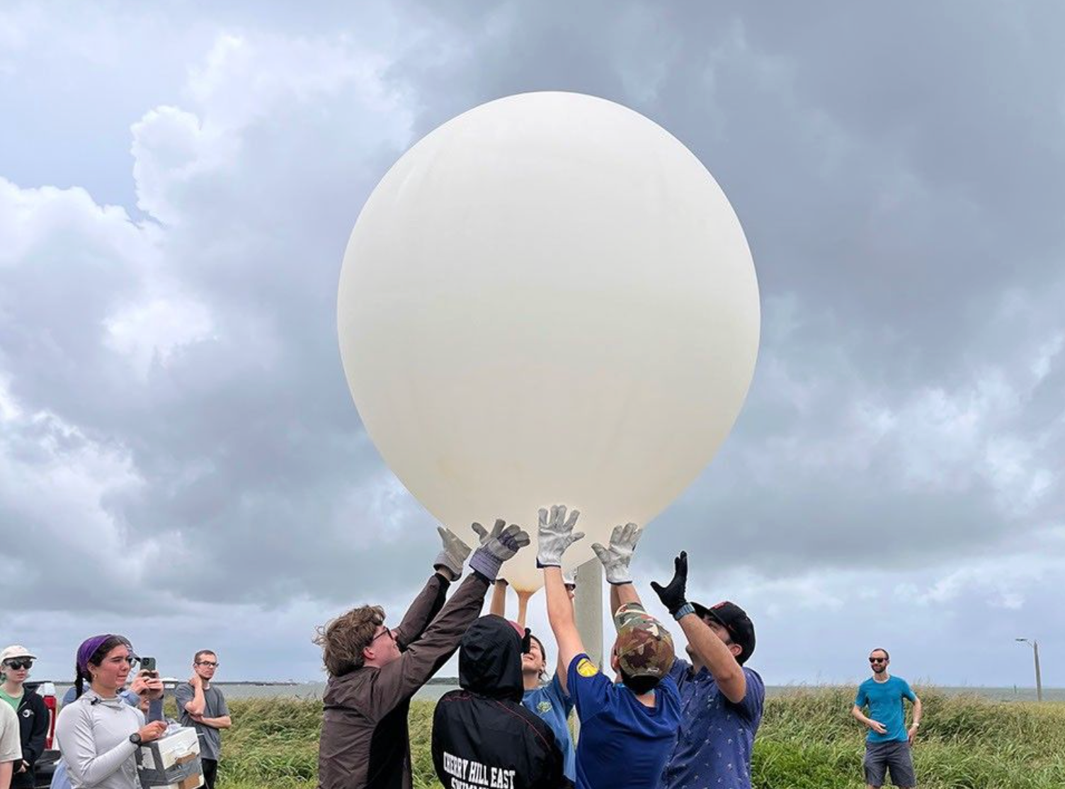 El centro de la imagen muestra un gran globo blanco. Debajo del globo, cinco personas forman un círculo y lo sostienen suavemente por la parte inferior para mantenerlo en posición vertical. Al fondo de la imagen, otras personas observan.