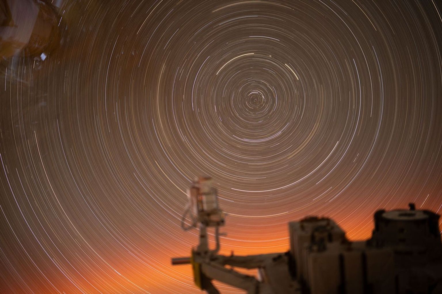 Esta fotografía de larga exposición, tomada durante 31 minutos desde una ventana del módulo laboratorio Kibo de la Estación Espacial Internacional, captura los arcos circulares de los rastros de las estrellas. En primer plano se ve una parte de la instalación expuesta del Kibo, donde se montan diversas cargas útiles y experimentos para exponerlos directamente al vacío del espacio.