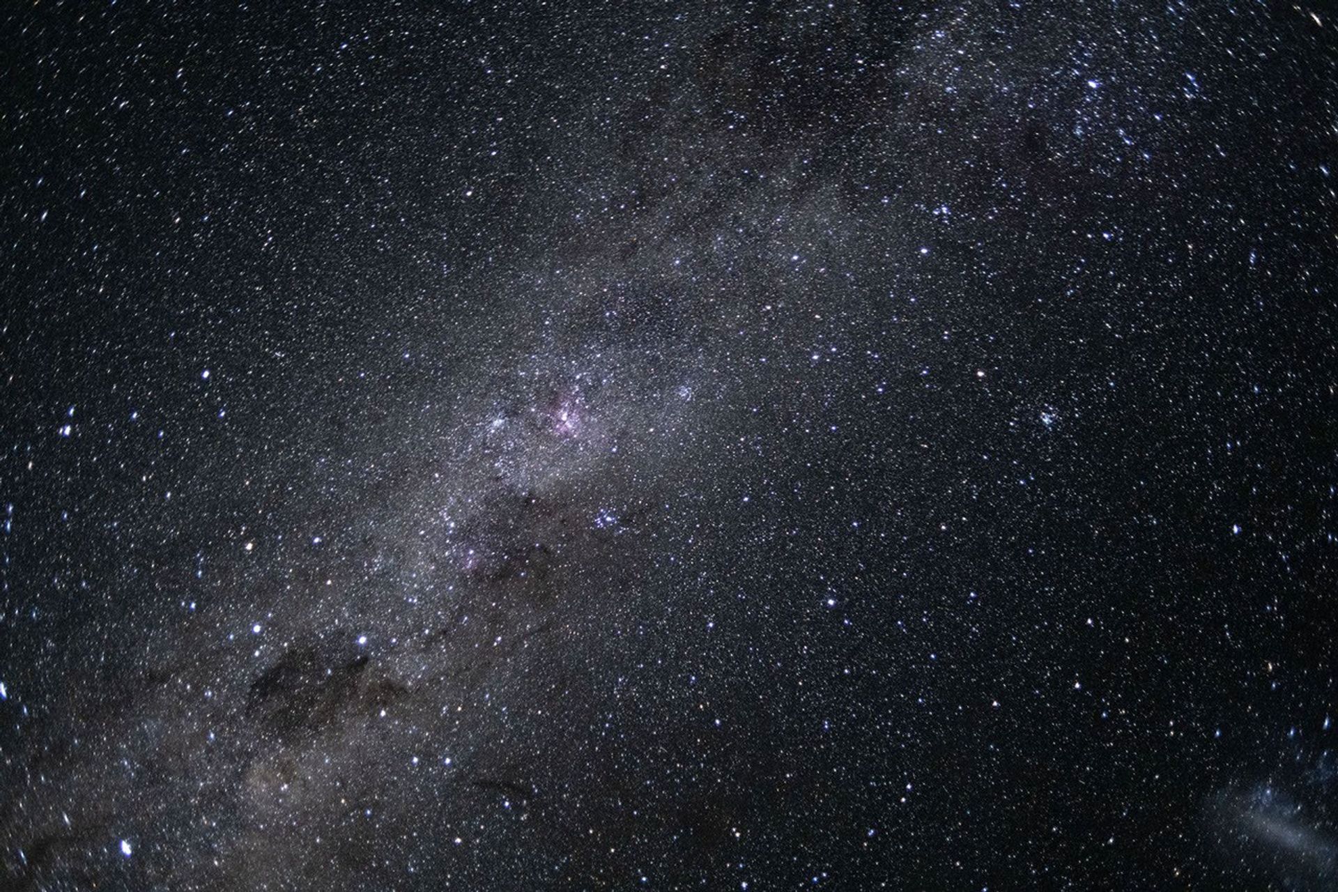 Un campo estelar repleto de miles de estrellas y resplandecientes nubes de polvo. El cielo detrás de las estrellas es negro. La Vía Láctea atraviesa el cielo en forma diagonal, formando una nube blanca y rosa que se extiende desde la parte inferior izquierda hasta la parte superior derecha.