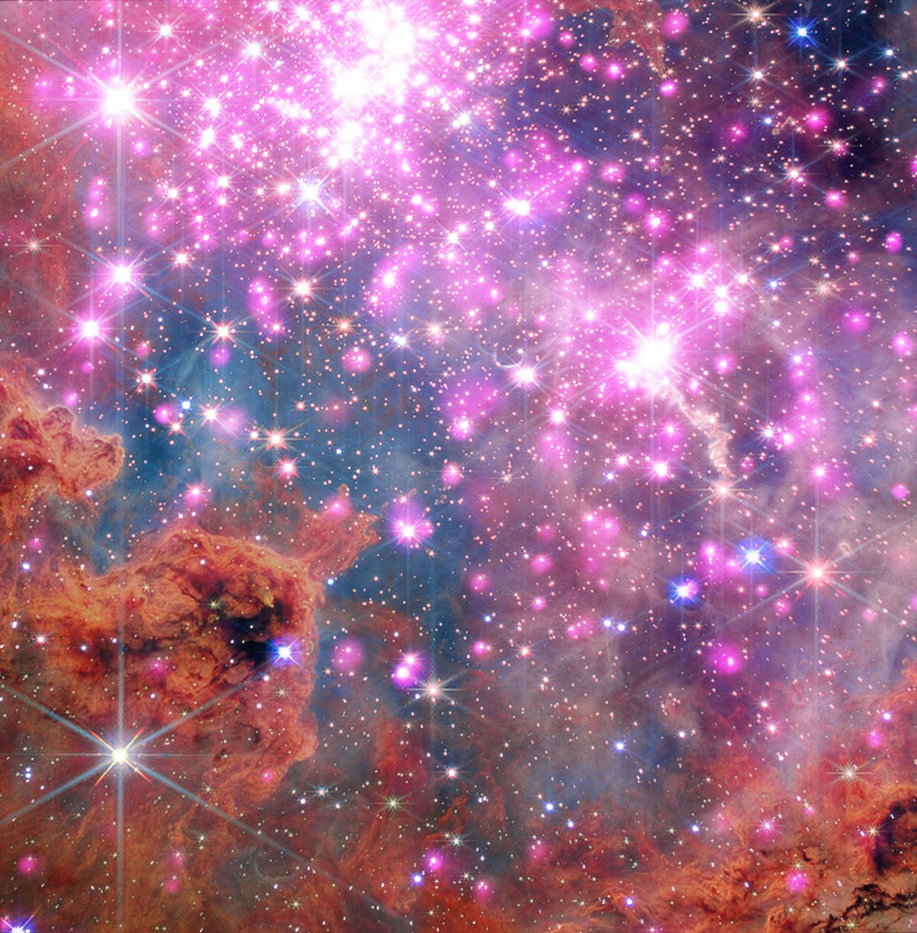 Esta imagen de Westerlund 2, publicada el 19 de marzo de 2026, presenta datos del Observatorio de rayos X Chandra (rosa) y datos infrarrojos del telescopio espacial James Webb de la NASA (rojo, naranja, verde, cian y azul). Decenas de estrellas resplandecientes rodeadas de rosa neón se extienden a lo largo de la imagen, destacando un cúmulo donde las estrellas tienen entre uno y tres millones de años de edad. Nubes de polvo color naranja ladrillo a lo largo del borde inferior ilustran las materias primas de esta activa guardería estelar.