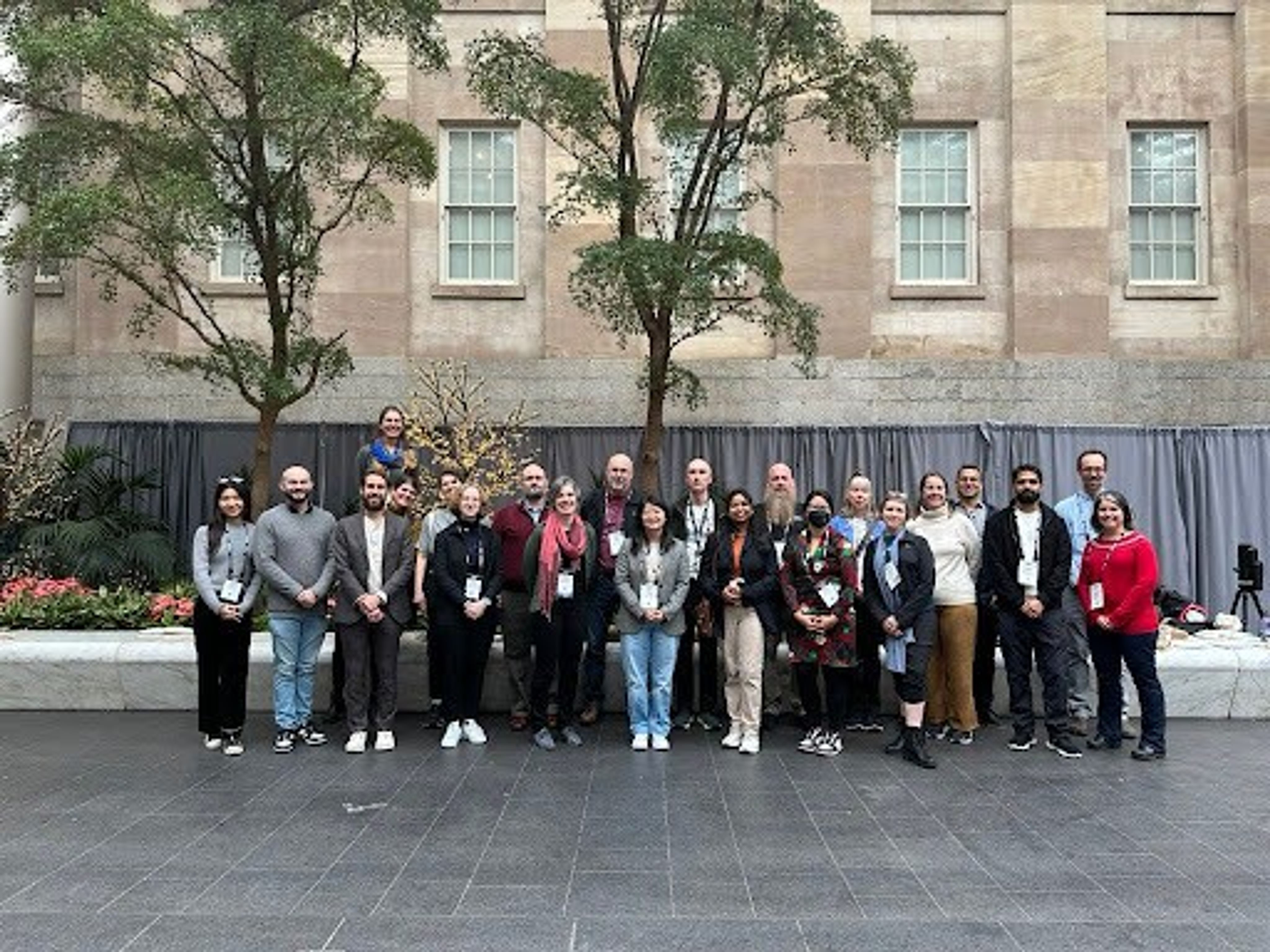 A group photo of attendees standing in a courtyard with tall trees and plants in the background. The group consists of diverse individuals dressed in casual and professional attire, posing in front of a stone building with large windows. The setting includes greenery, floral arrangements, and a paved floor, suggesting a formal event or conference.