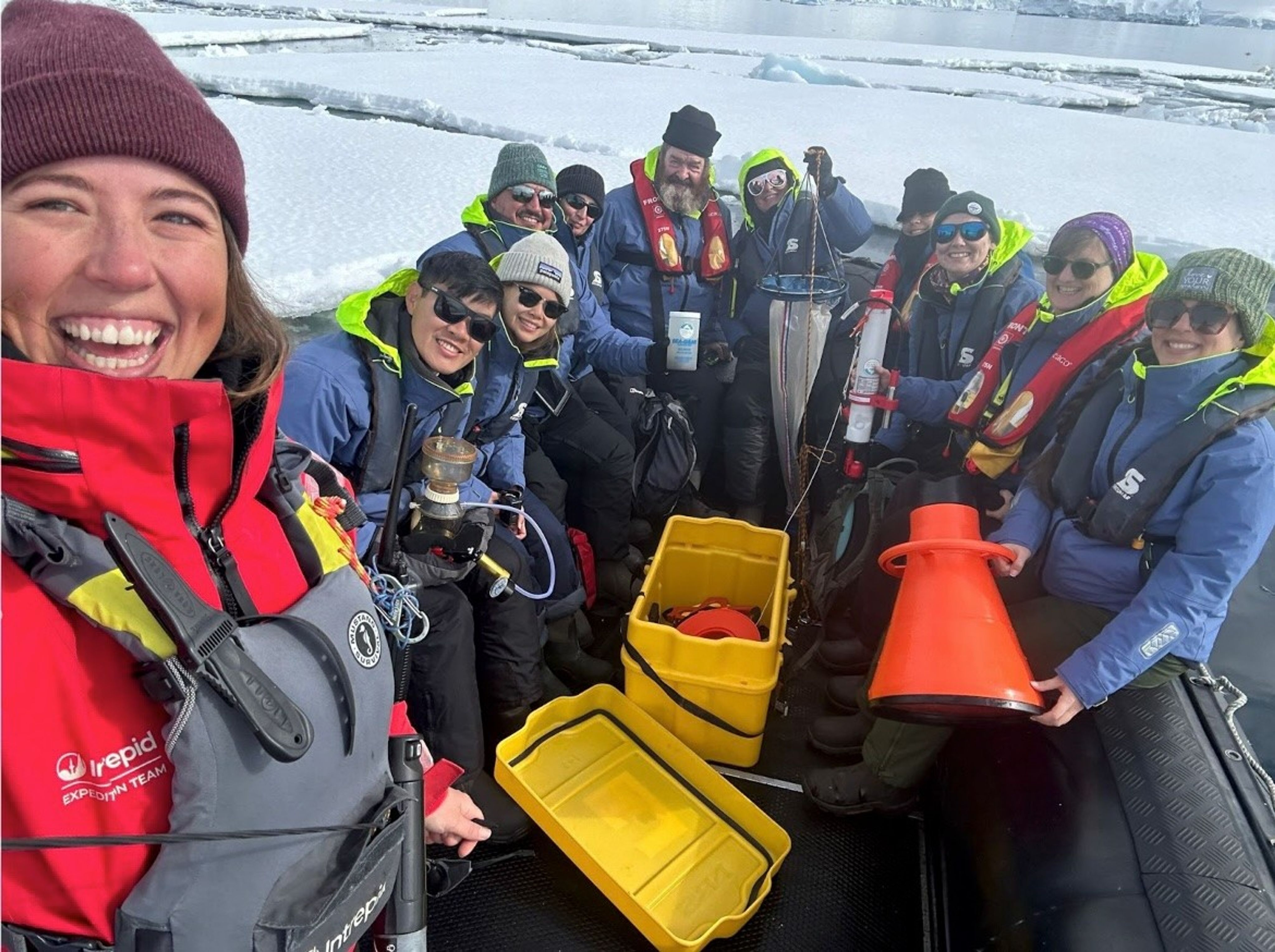 A group of smiling people in cold-weather gear and life jackets pose on a boat surrounded by icy waters. They are holding scientific equipment, including a plankton net and collection bottles, with yellow bins and an orange traffic cone visible in the foreground.