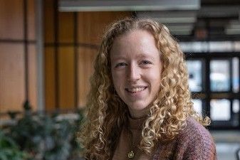 A young woman with curly blonde hair wearing a cozy sweater smiles at the camera, standing in a modern hallway with plants in the background.