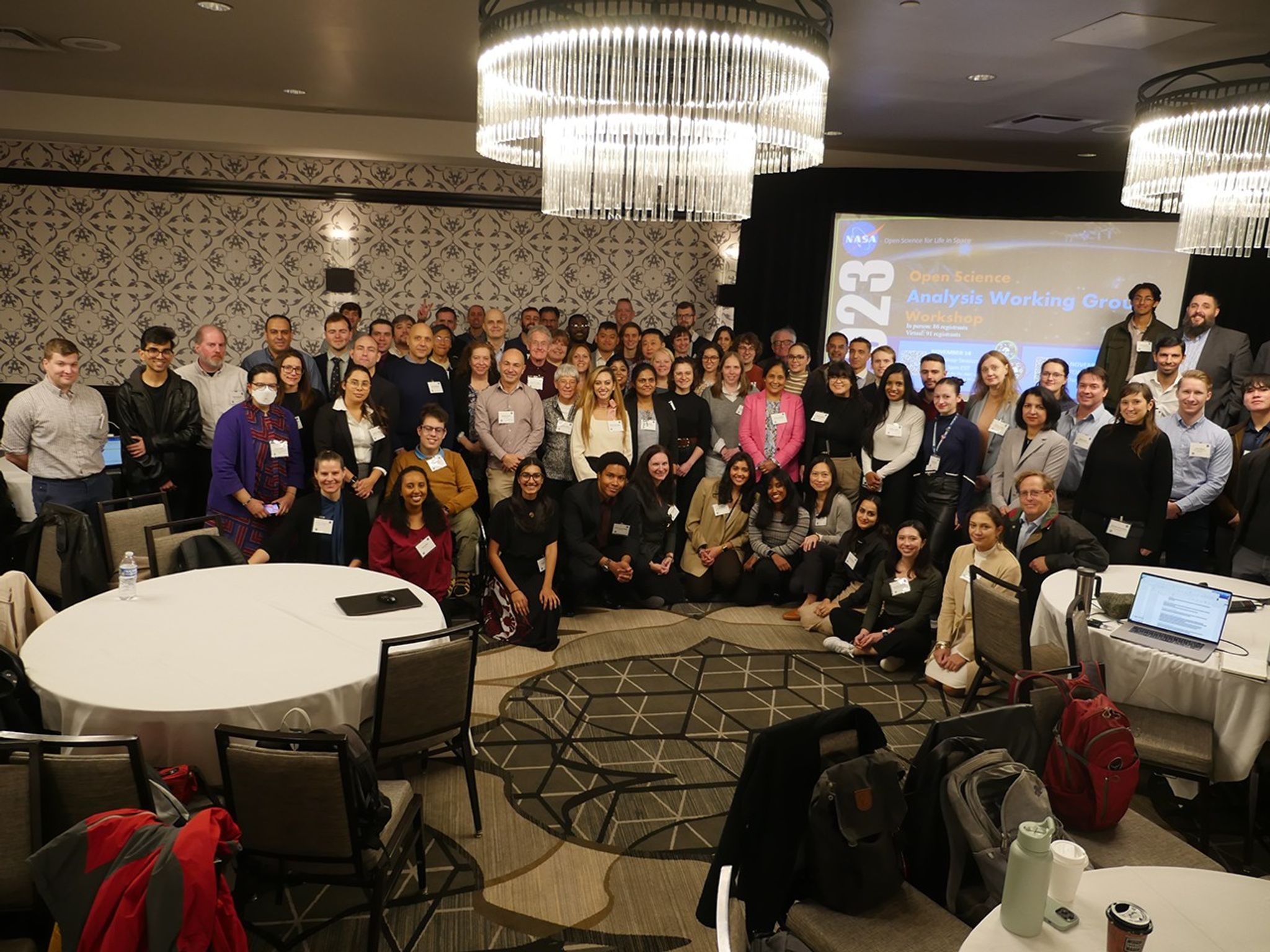 Large group photo of attendees at a NASA Analysis Working Group meeting, gathered in a conference room with round tables and chandeliers. A presentation screen is visible in the background.