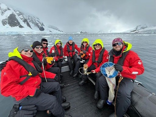 A group of eight people wearing red and black cold-weather gear with neon yellow hoods are seated in a black inflatable boat in a polar environment. They are smiling and holding scientific equipment, including a plankton net, a mug, and an orange device. Snow-covered mountains and an icy ocean surround them under an overcast sky.