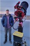 Man standing next to a large red telescope on an outdoor observatory platform at sunset.