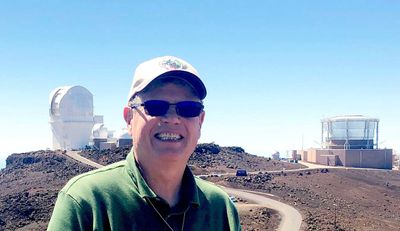 A man wearing a green polo shirt, sunglasses, and a baseball cap smiles at the camera while standing outdoors in a rocky, high-altitude location. Behind him, there are observatory buildings with large dome-shaped and cylindrical structures. The sky is clear and bright blue.