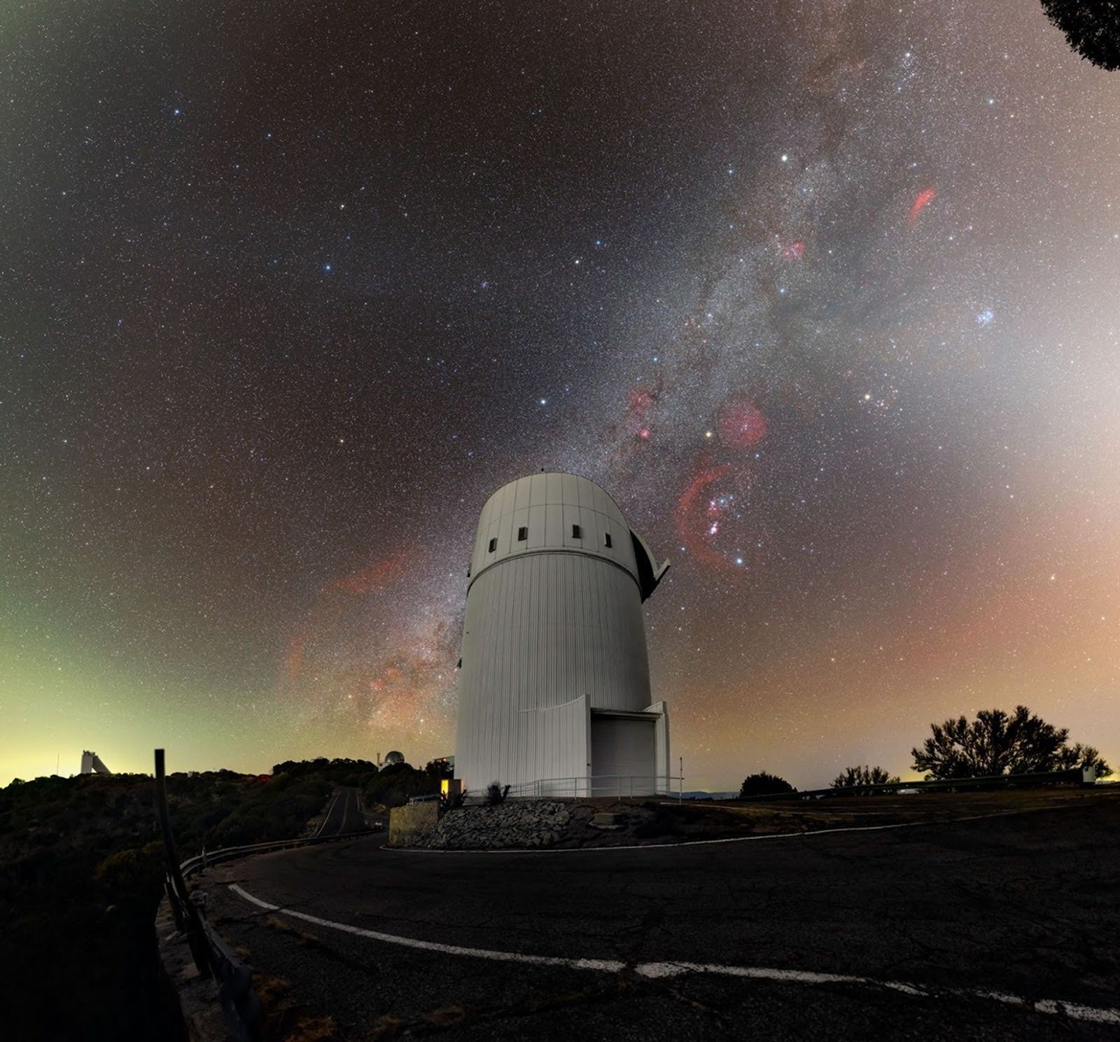 Nighttime view of the white Bok telescope dome at Kitt Peak National Observatory beneath a star-filled sky. The Milky Way stretches overhead with dense star clouds and reddish nebulae visible, while the observatory sits beside a curved road on a dark hillside.
