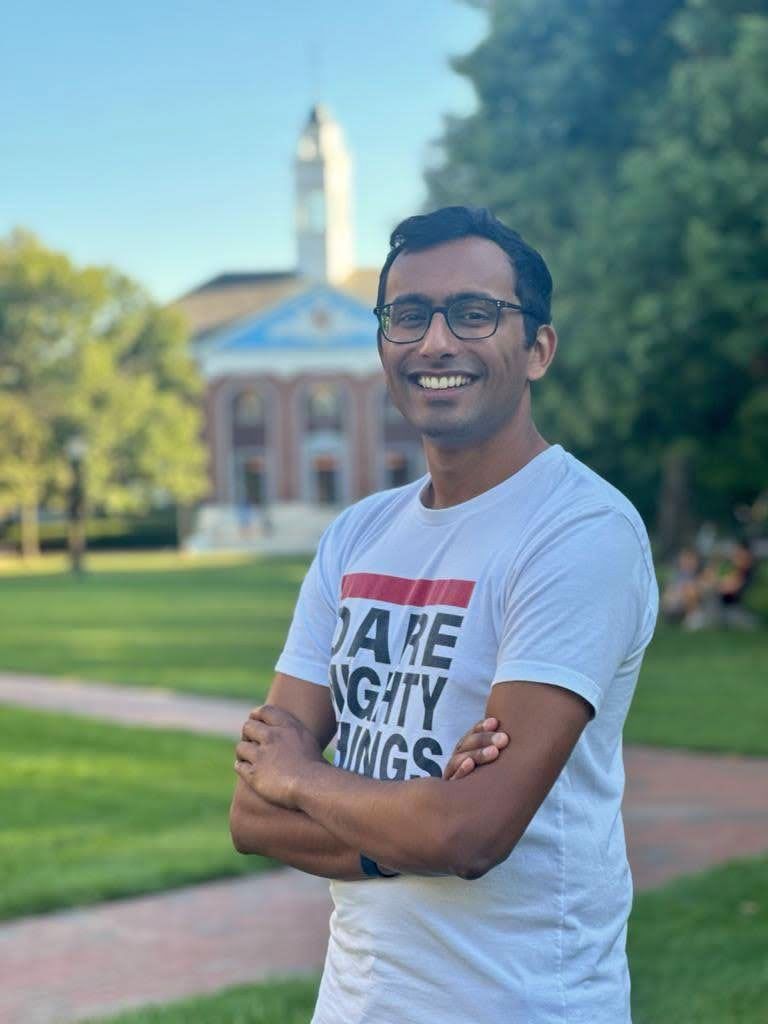 Portrait of Ben Fernando wearing glasses and a t-shirt standing outdoors with arms crossed in front of a campus-style building and green lawn.
