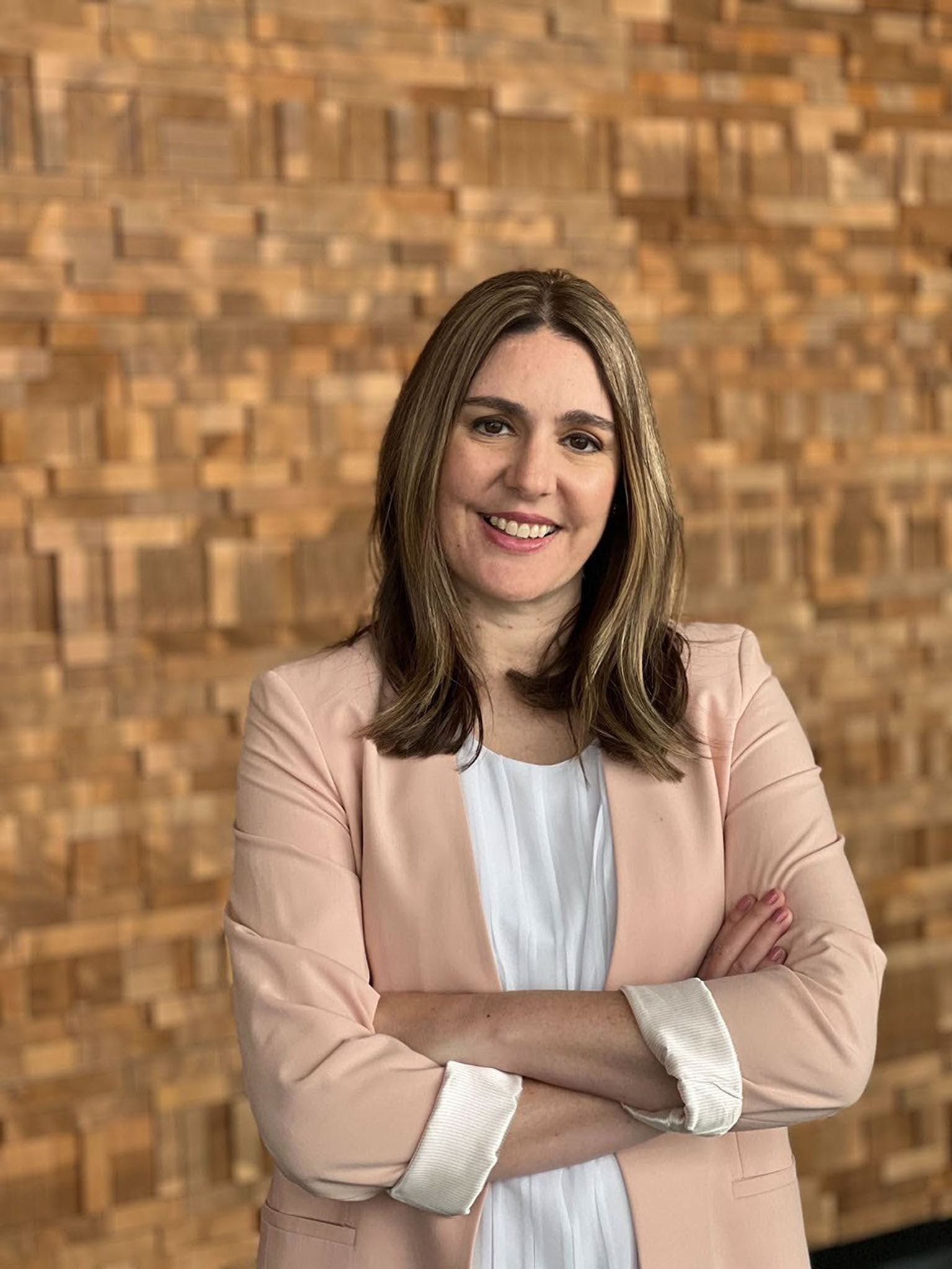 Headshot of Jennifer Heldmann standing with arms crossed, wearing a light‑colored jacket and white top, in front of a textured wooden wall.