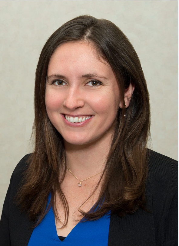 Headshot of Kelsey Young wearing a blue top and dark jacket, posed against a plain light-colored background.