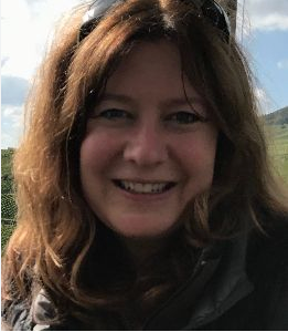 Headshot of Maria Banks outdoors with sunlight and a natural landscape and sky in the background.