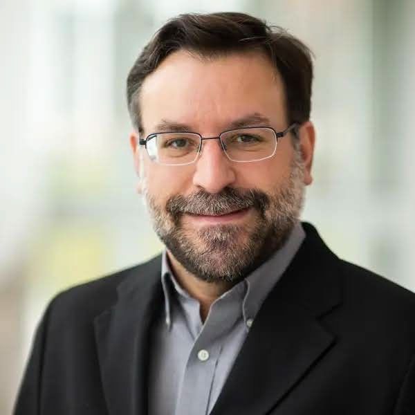 Headshot of Nicholas Schmerr wearing a collared shirt and dark jacket, indoors, with a softly blurred light-colored background.