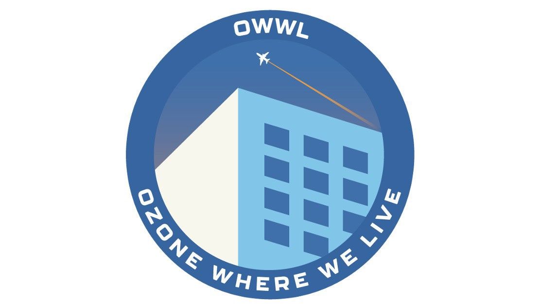 A circular logo with a blue background. The acronym "OWWL" sits at the top. Inside the circle is a stylized light blue building, with a small white airplane flying diagonally above it, leaving an orange trail. The words "OZONE WHERE WE LIVE" are written along the bottom curve of the circle.