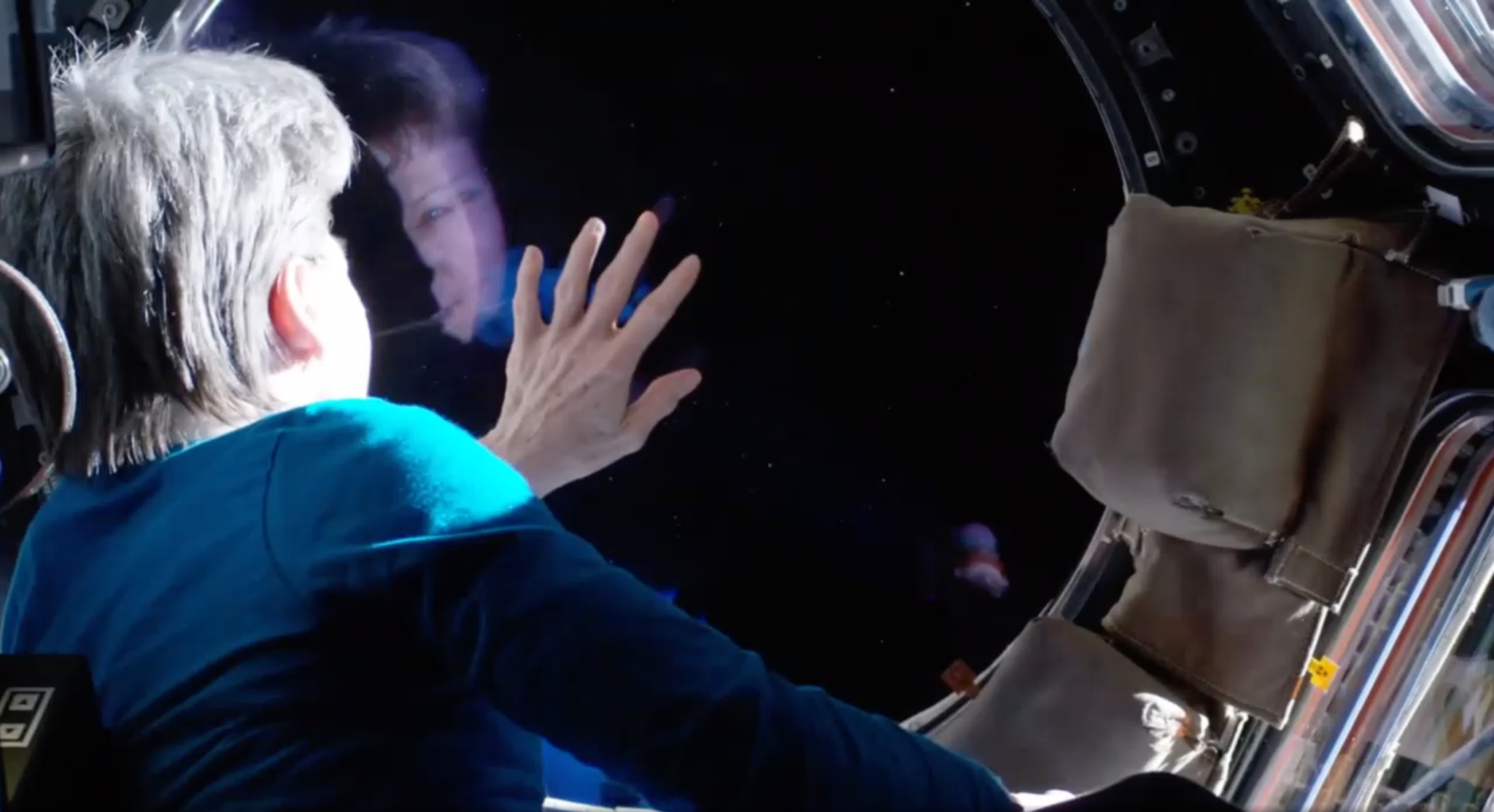 Inside the International Space Station’s Cupola, an astronaut with short, graying hair wearing a blue shirt leans forward and presses her outstretched hand against a large circular window. Her face is partially reflected in the glass. She looks out into the dark expanse of space, dotted with faint stars. A tan storage pouch is attached to the right side of the module’s interior.