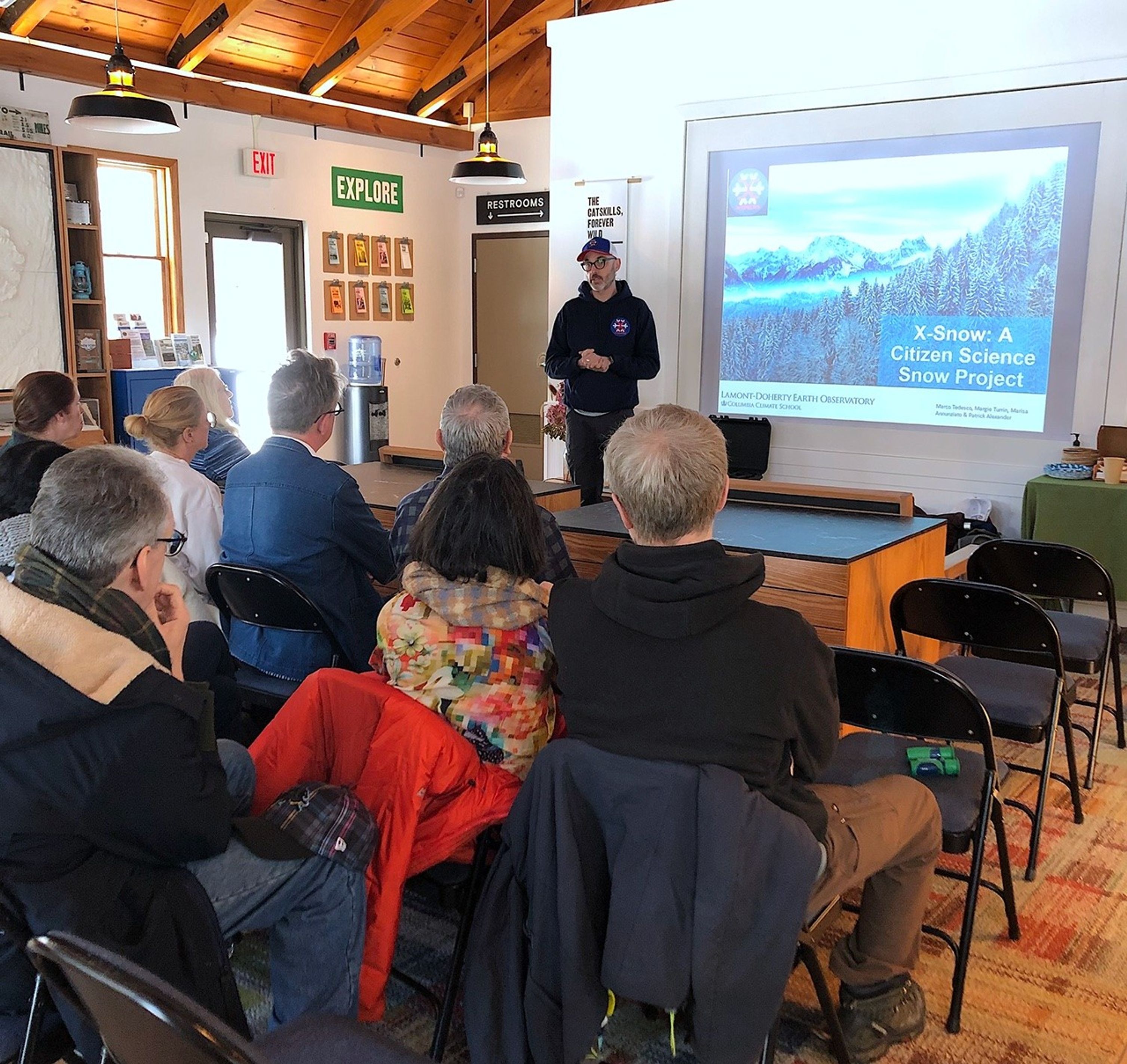 A man in a baseball hat stands next to a projected image of snow-dusted evergreen trees with snow-capped peaks in the background. Eight mostly grey-haired people sit in chairs facing the man, their colorful winter coats draped over their chair backs.