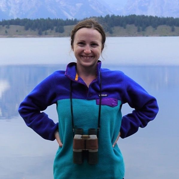 Person smiling with hands on hips, wearing a blue and teal fleece and binoculars around their neck, standing in front of a calm lake with mountains in the background.