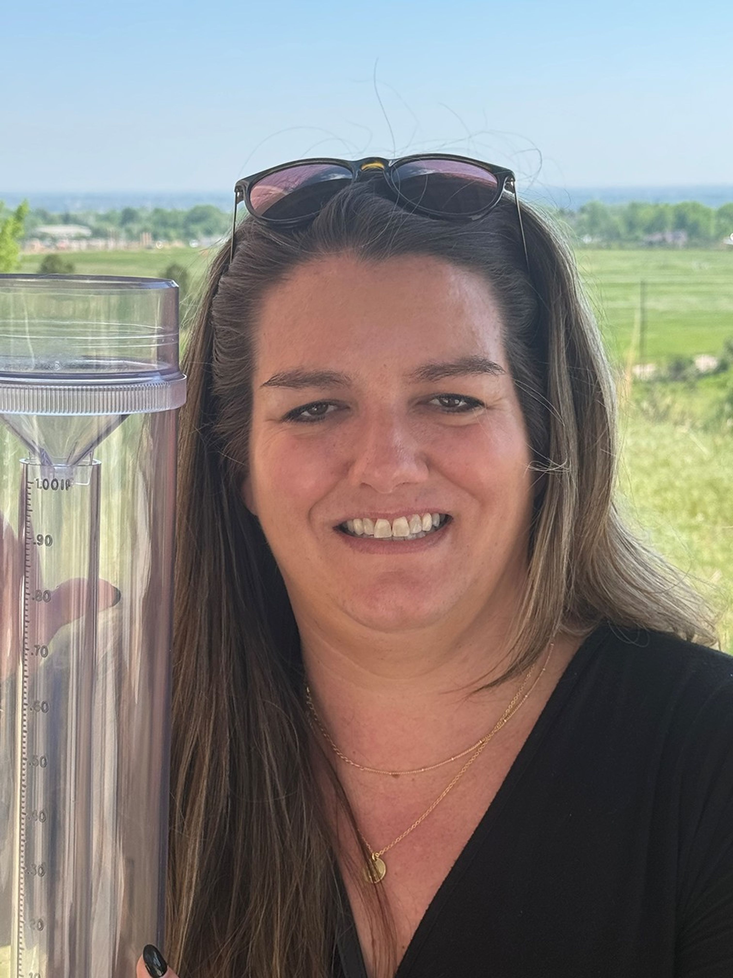 White woman in dark v-neck shirt holds up a rain and snow gauge.