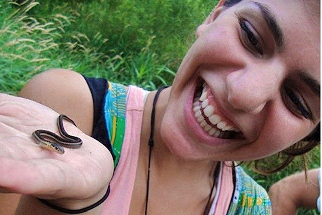 Smiling person outdoors holding a tiny snake in their hand, looking closely at it with excitement.