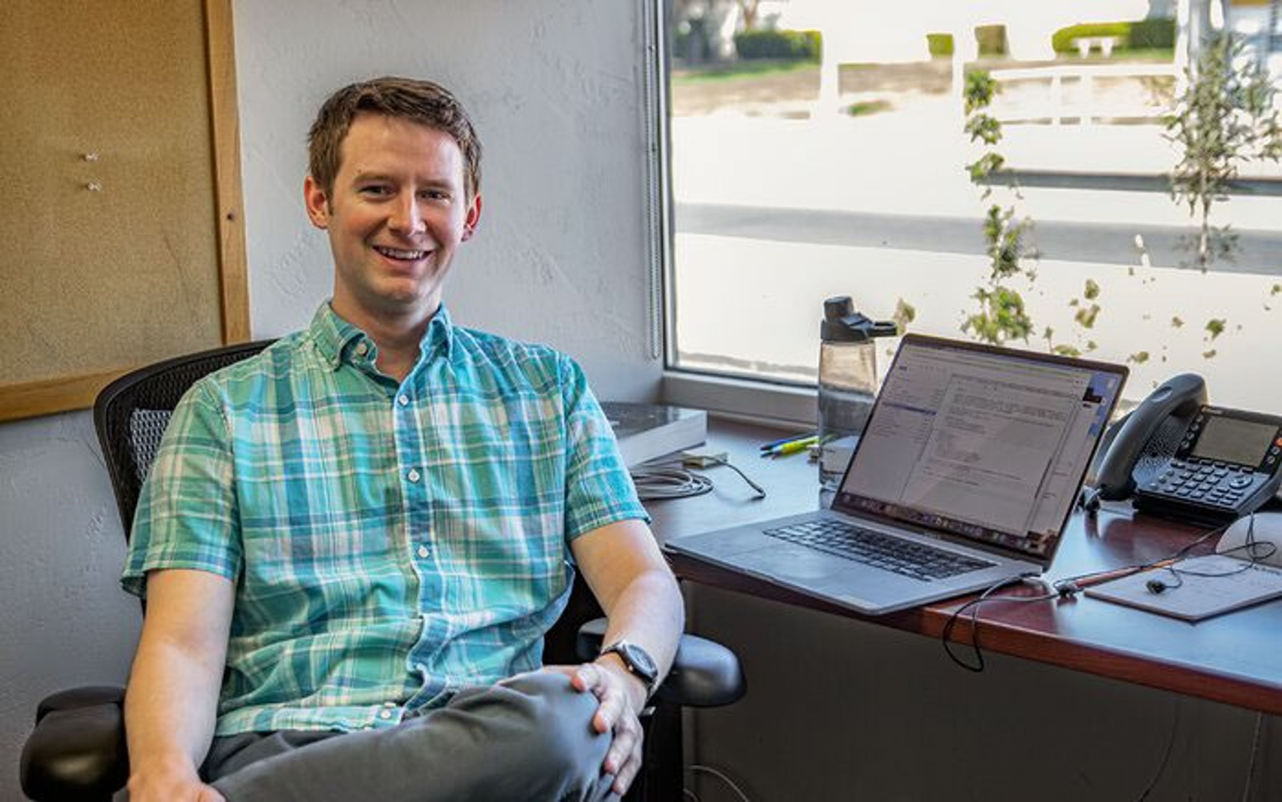 Photo of a young smiling man sitting at a desk with a laptop