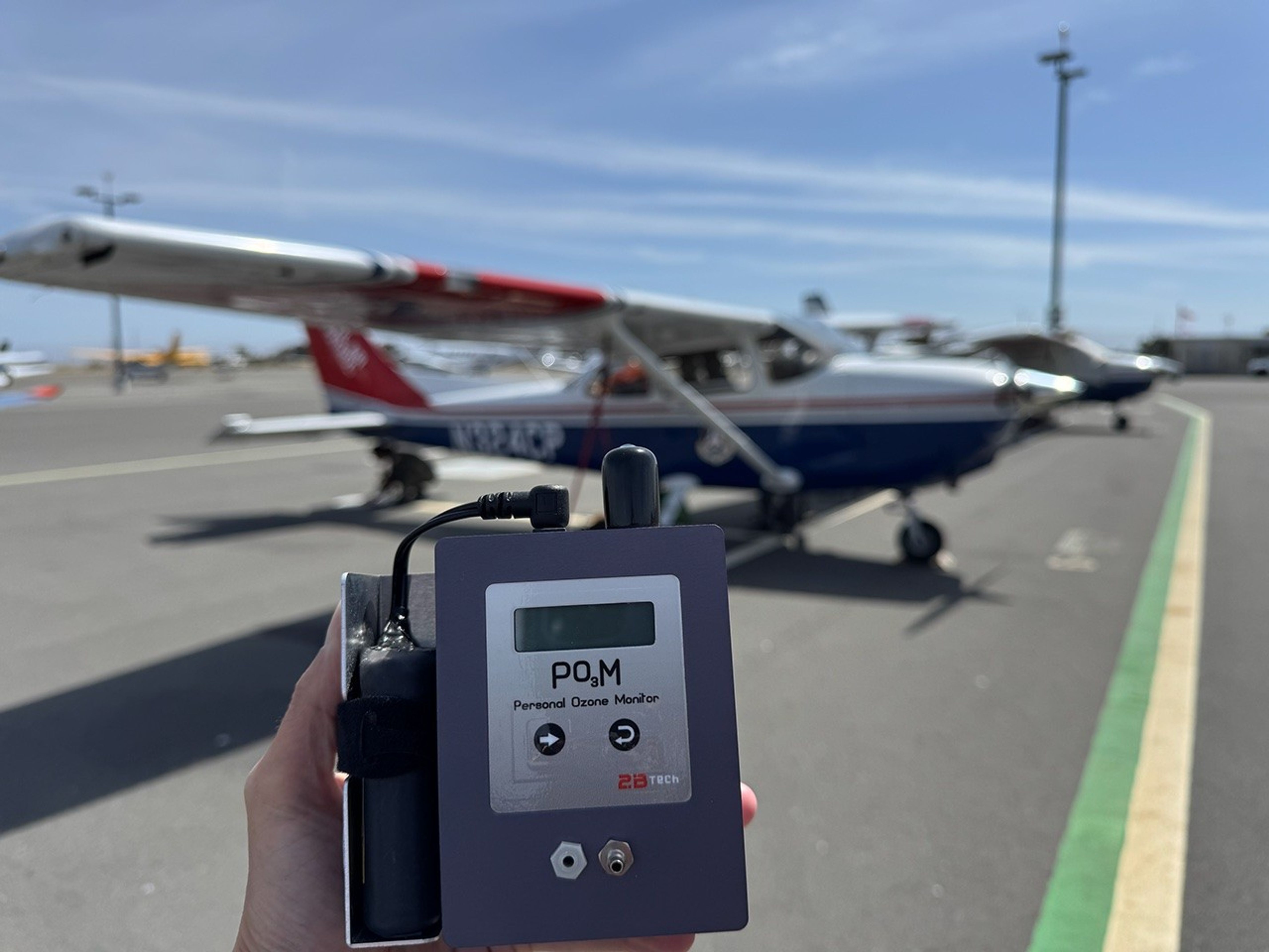 A hand holds a grey control box with plugged in components with the words “Personal Ozone Monitor” on its front. In the background is a small fixed-wing aircraft.