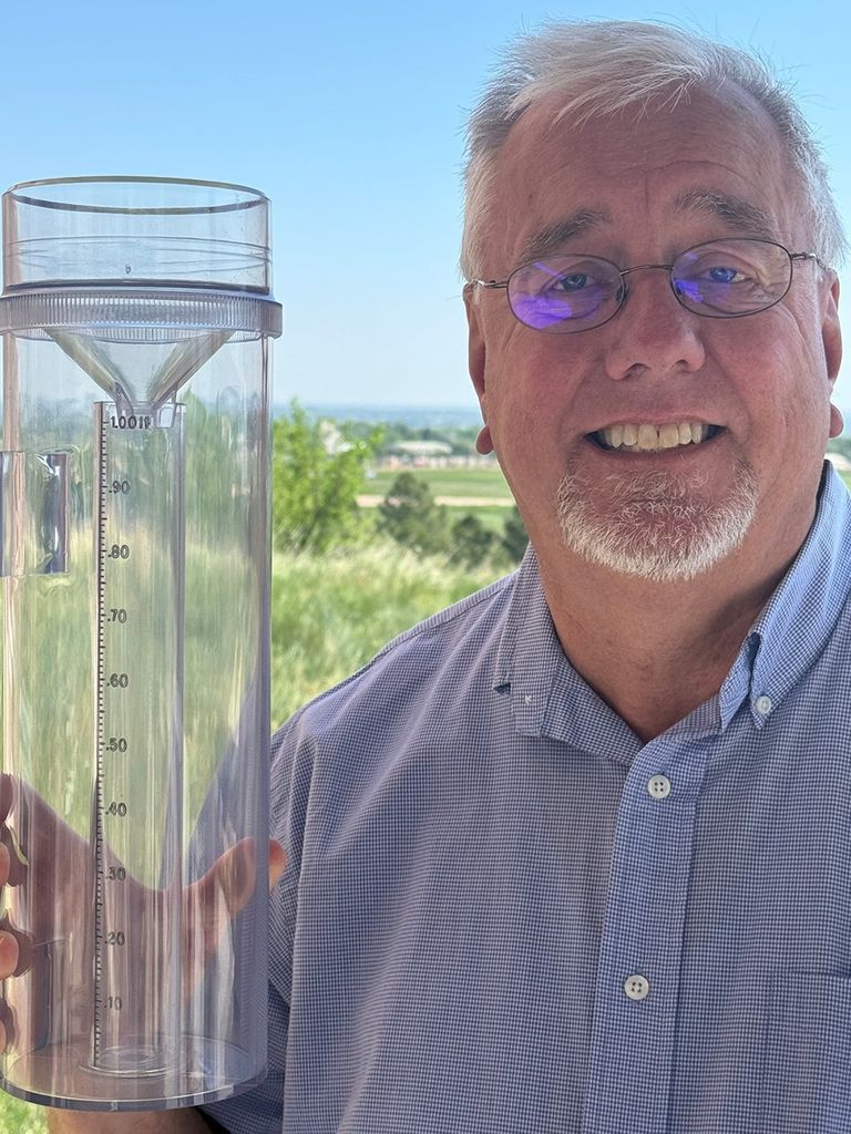 White man in blue shirt holds up a rain and snow gauge.