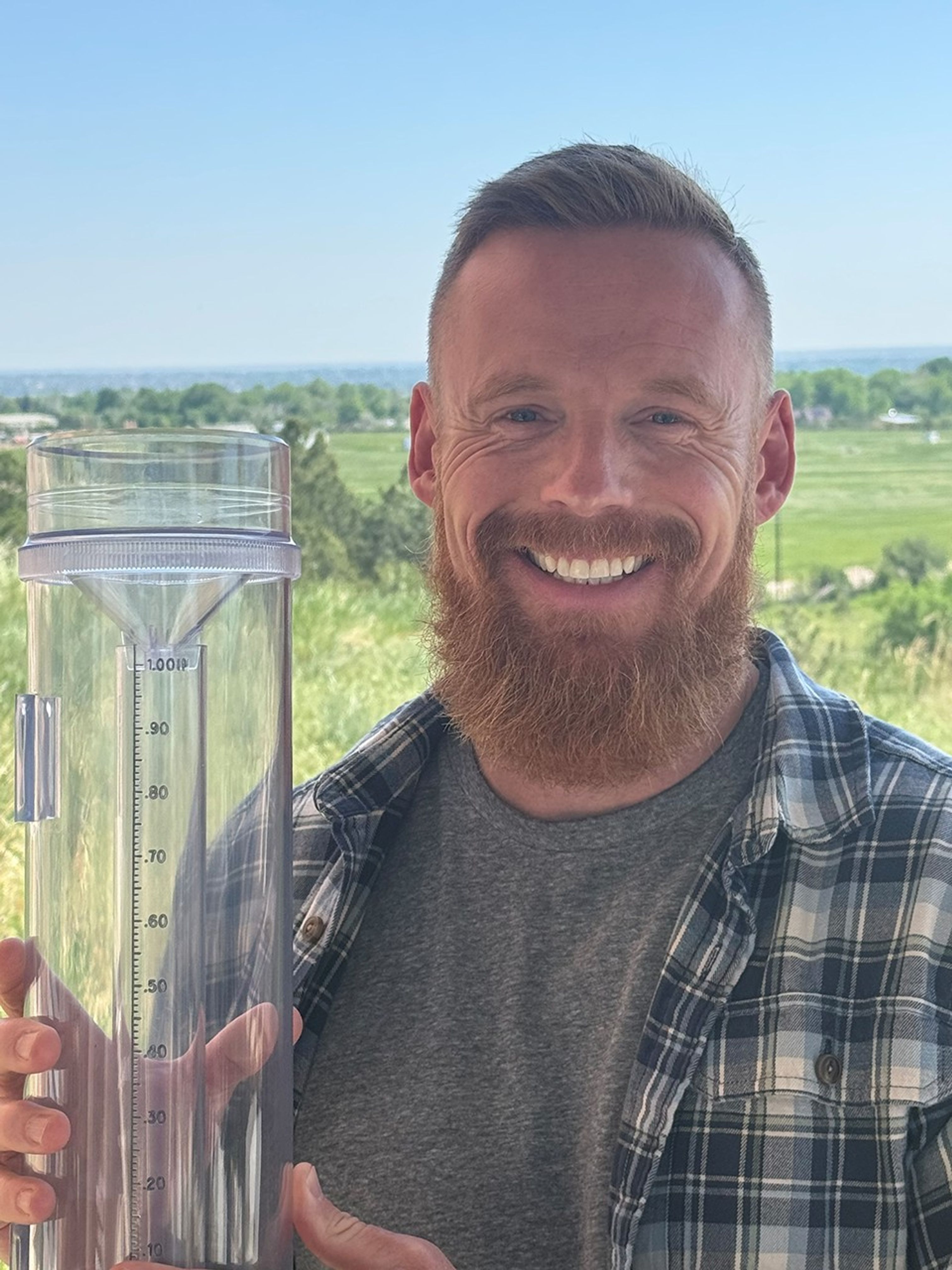 White man in blue and white plaid shirt holds up a rain and snow gauge with a big smile on his face.