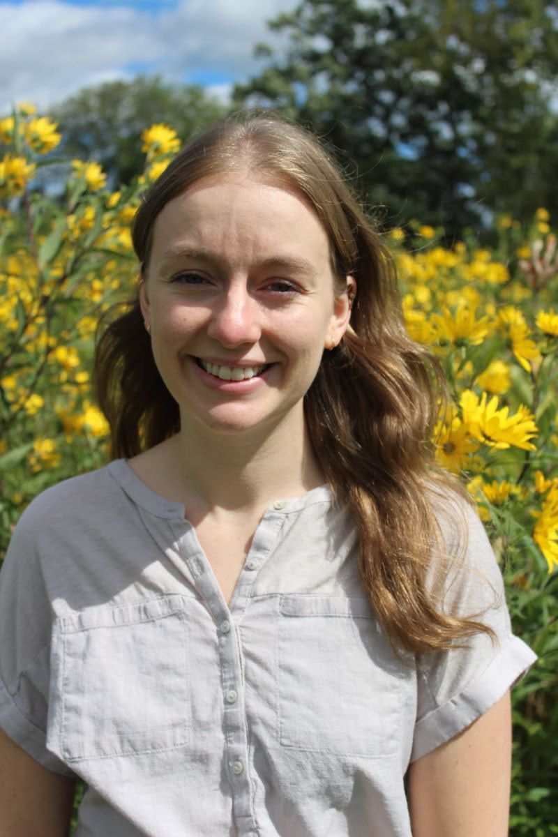 Photo of a young smiling woman standing in front of a field of yellow flowers