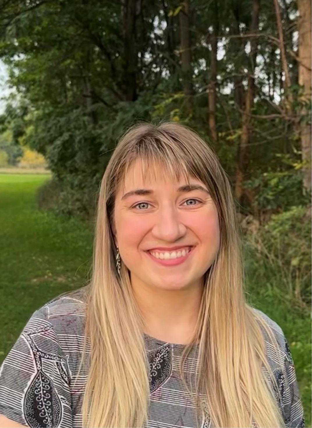 Photo of a young smiling woman with lush green trees and grass in the background