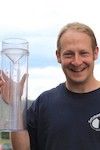 A white man in black tee shirt holds up a rain and snow gauge with a big smile on his face.
