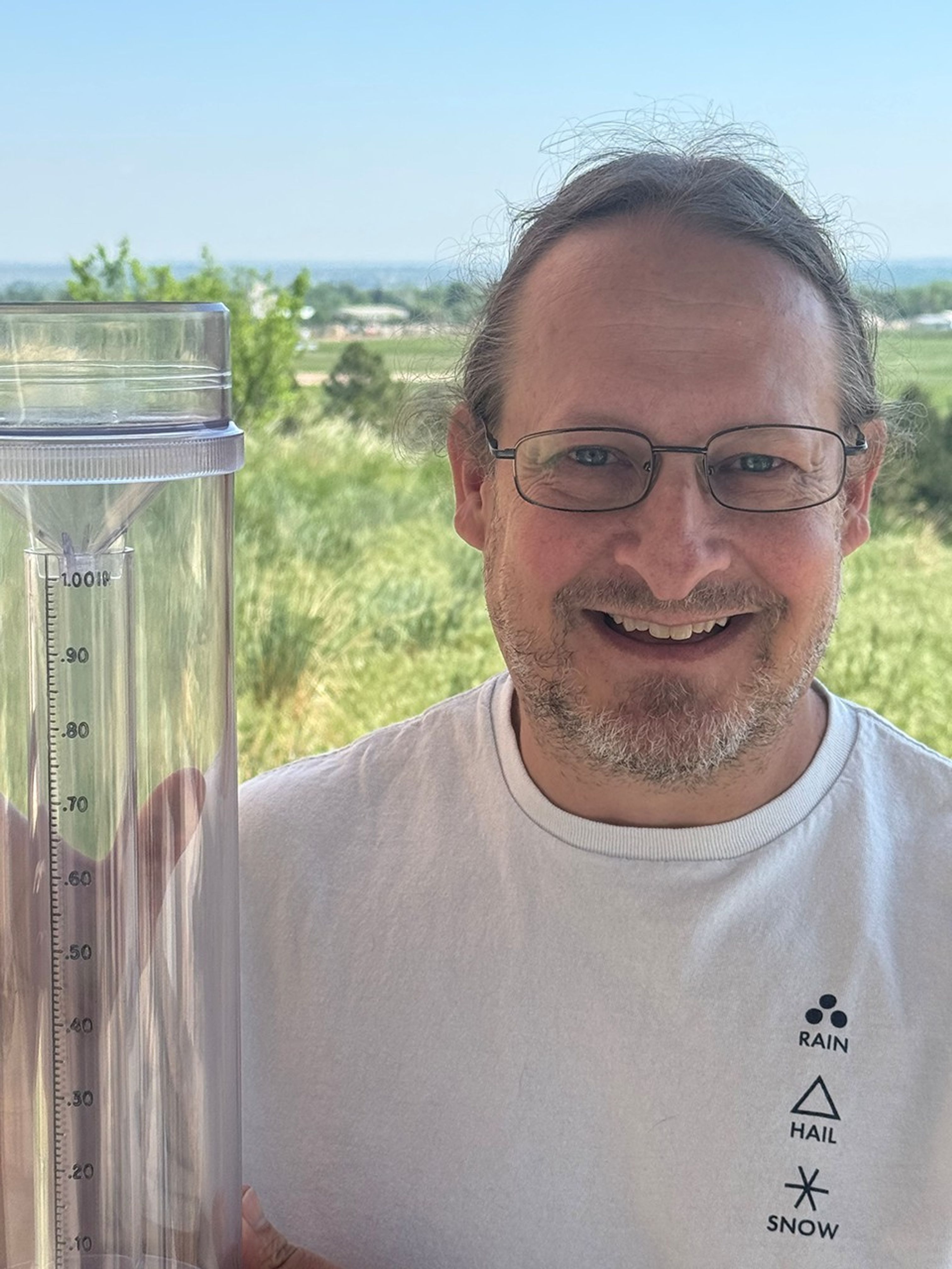 A white man in white tee shirt holds up a rain and snow gauge with a smile on his face.