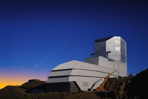 The boot-shaped building that houses the Vera C. Rubin Observatory shines pearly grey against a dawn or dusk sky. The faint streak of a comet is visible in the blue, just over the golden glow of the Sun just over the horizon. A yellow construction crane sits next to the building.