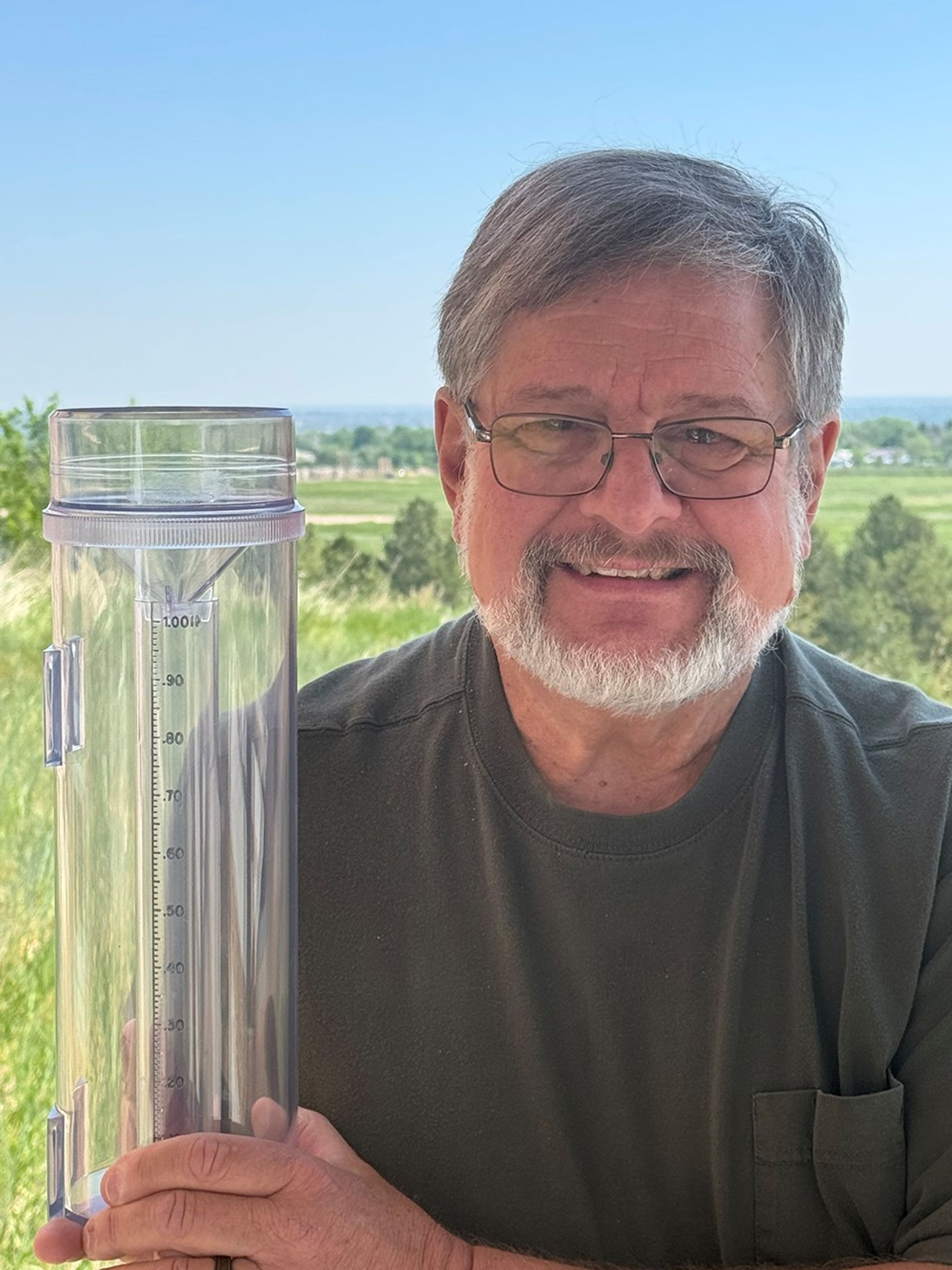 A white man with beard and dark tee shirt holds up a rain and snow gauge.