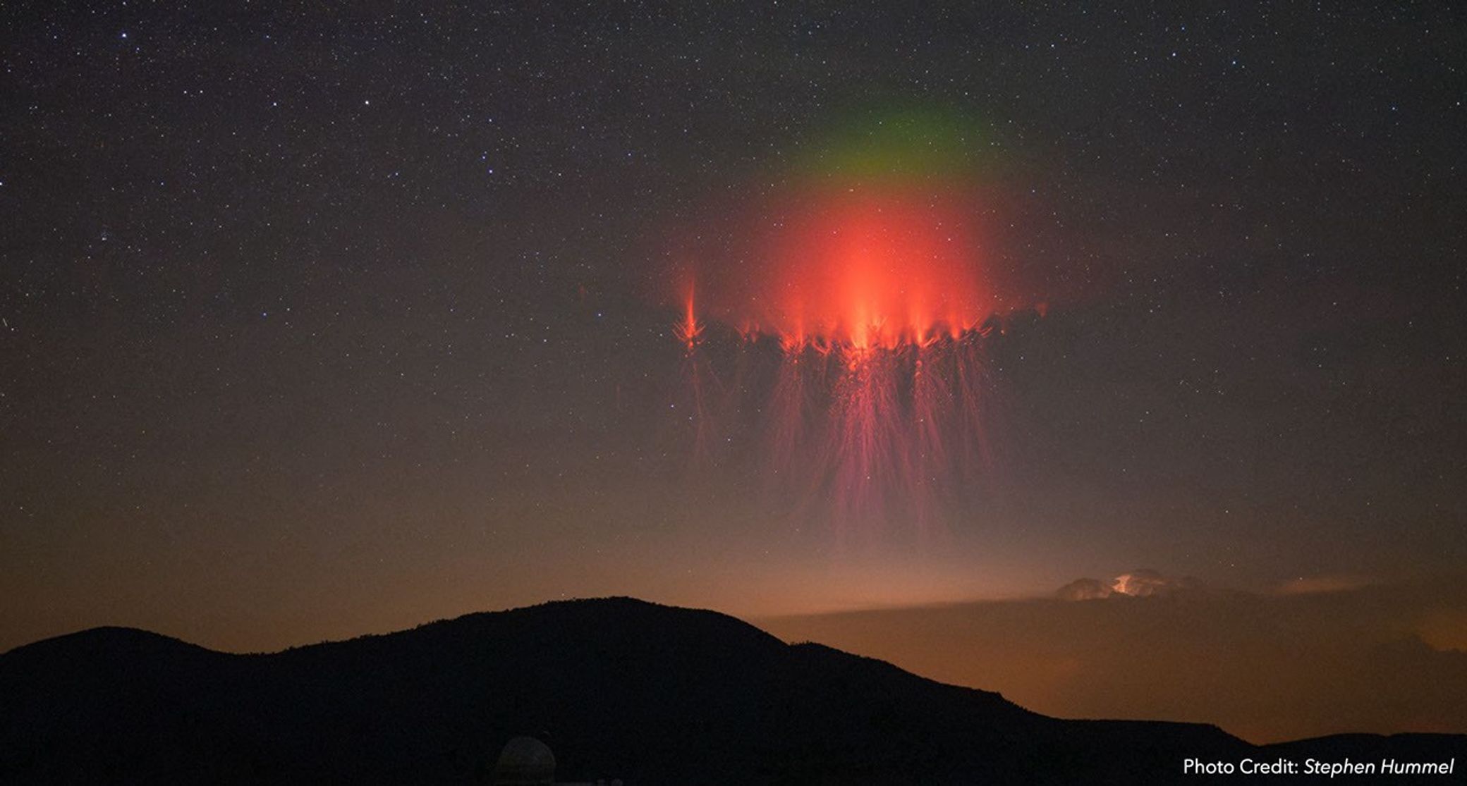 A vivid red sprite lightning burst appears high in the night sky above a distant mountain range. The sprite glows with intricate, tendril-like patterns and is topped with a faint green glow, set against a starry background.