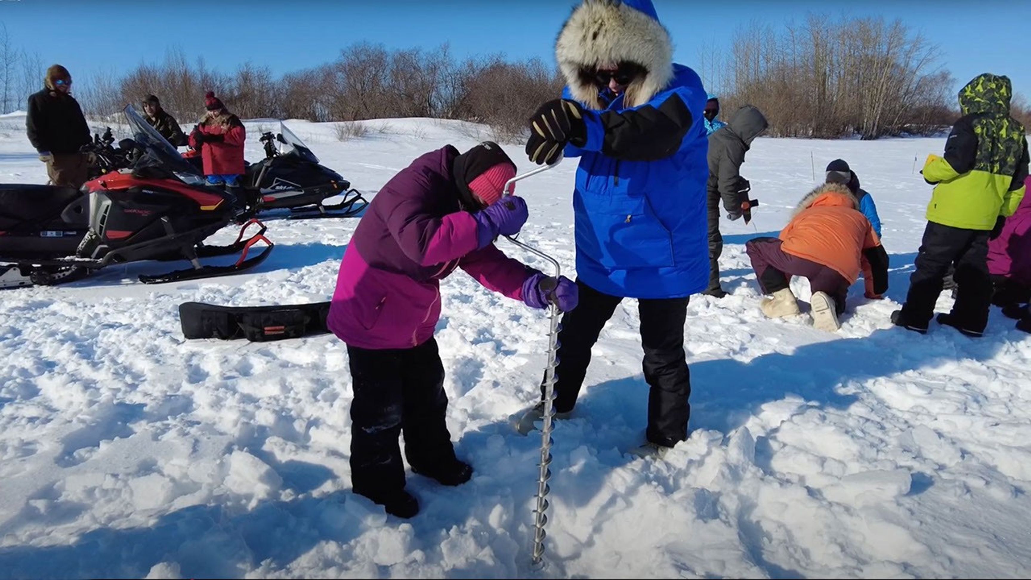 People bundled in winter gear drill a hole into the ice with a hand auger during an ice fishing outing, with snowmobiles and other participants in the background.