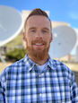 White man in blue green white plaid shirt in front of large satellite dishes