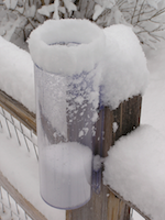 CoCoRaHS rain gauge, which looks like a skinny clear plastic tube inside a bigger clear plastic tube, attached to a weathered wooden post. Both post and gauge, inside and out, have a layer of snow.