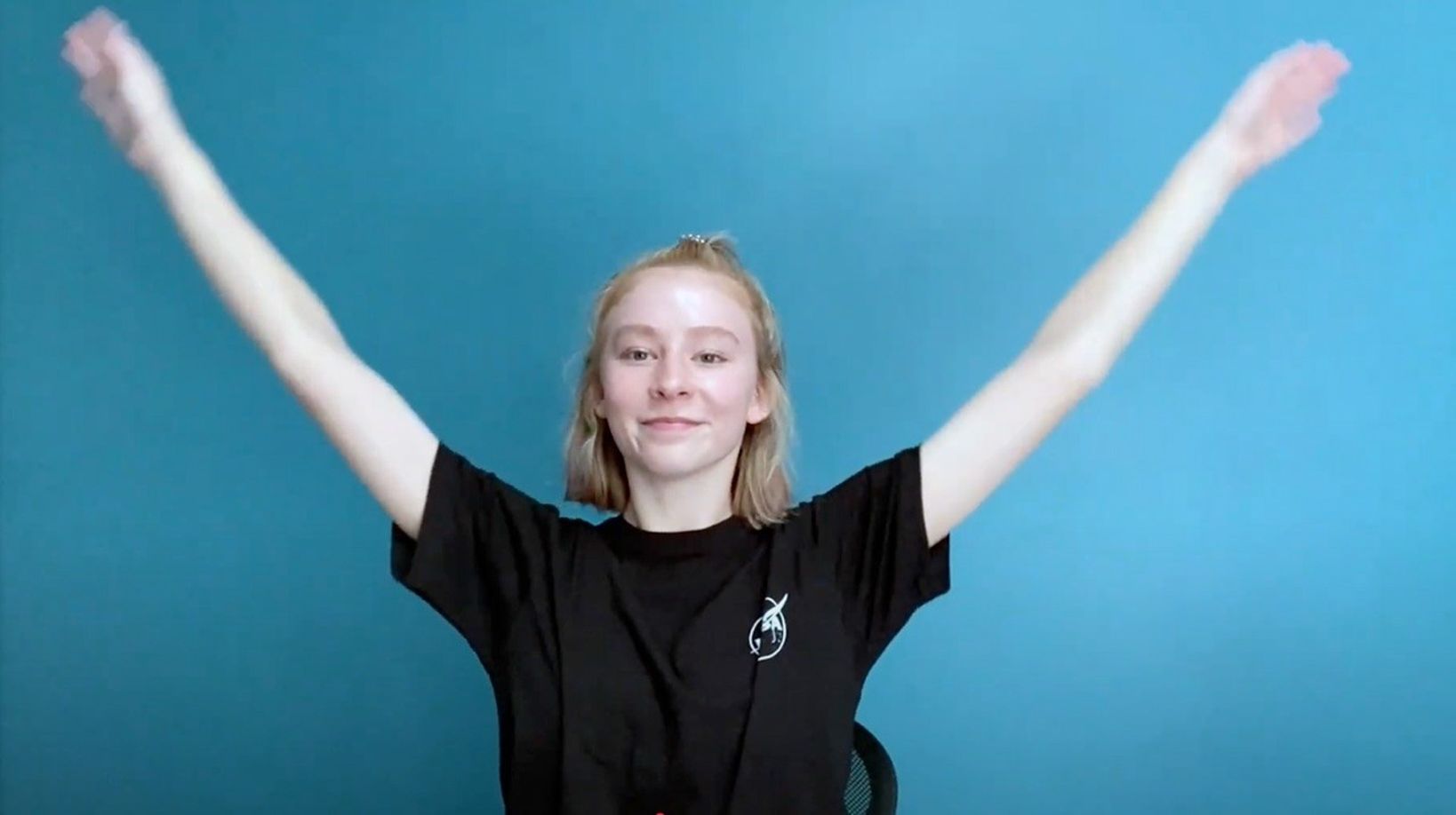 Student with arms outstretched, mid-pose against a blue background, wearing a black shirt with a space-themed logo.