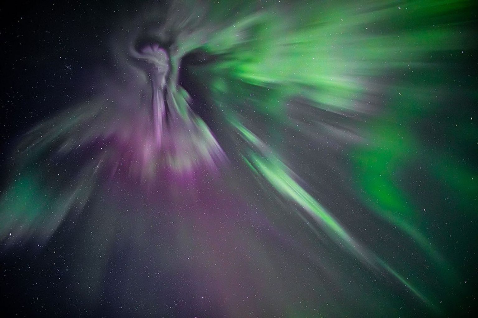 Stunning image of the aurora borealis in the night sky, featuring vibrant green and purple lights spreading out in dynamic, wispy patterns. The lights appear to converge at a central point, resembling a celestial explosion or ethereal figure, against a backdrop of countless stars.