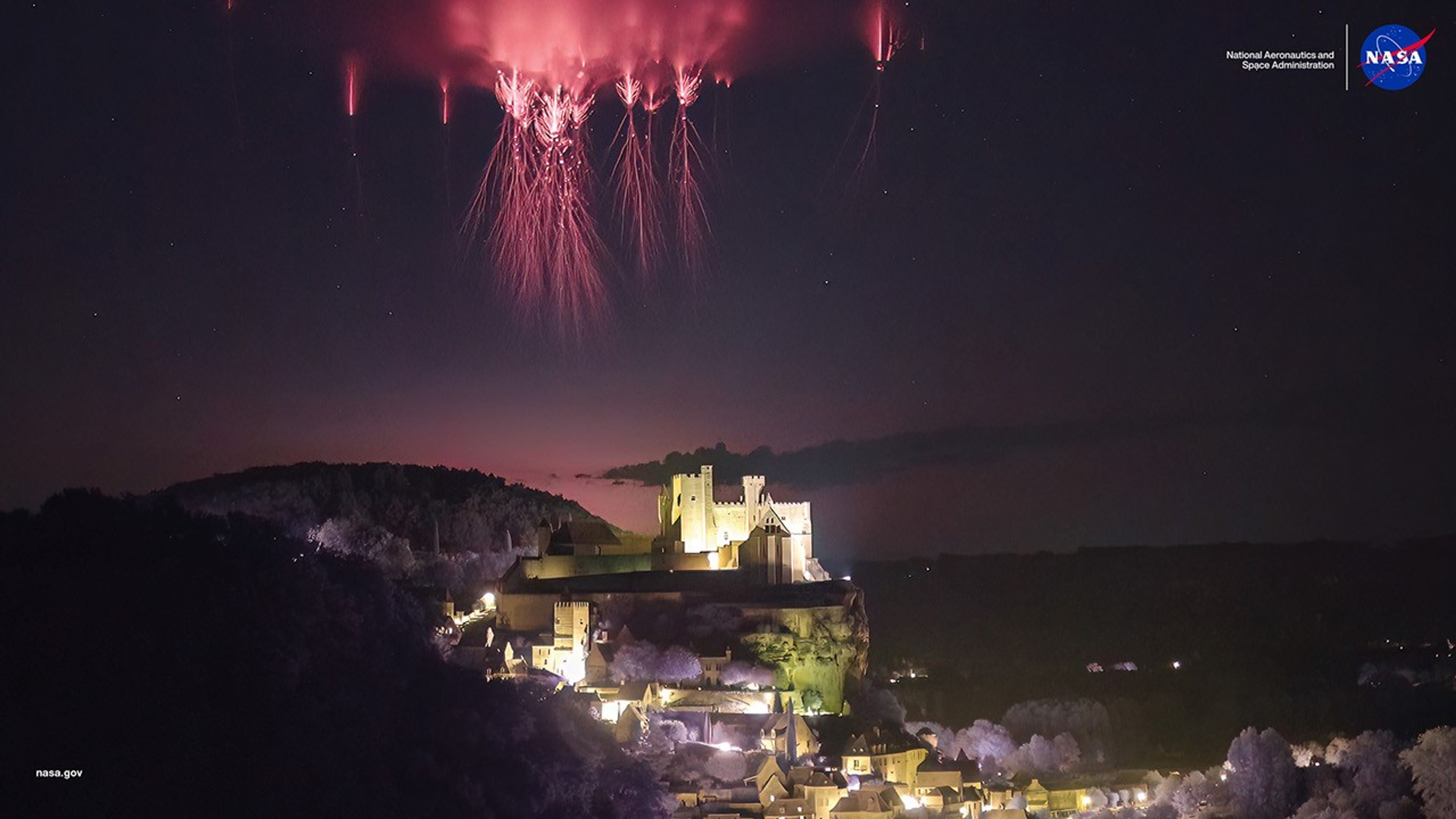 Nighttime photograph showing a red sprite, a rare upper atmospheric electrical phenomenon, appearing as a crimson flash above Château de Beynac castle in Dordogne, France. The sprite extends vertically in the dark sky above storm clouds, captured by NASA Spritacular citizen science project participant Nicolas Escurat.