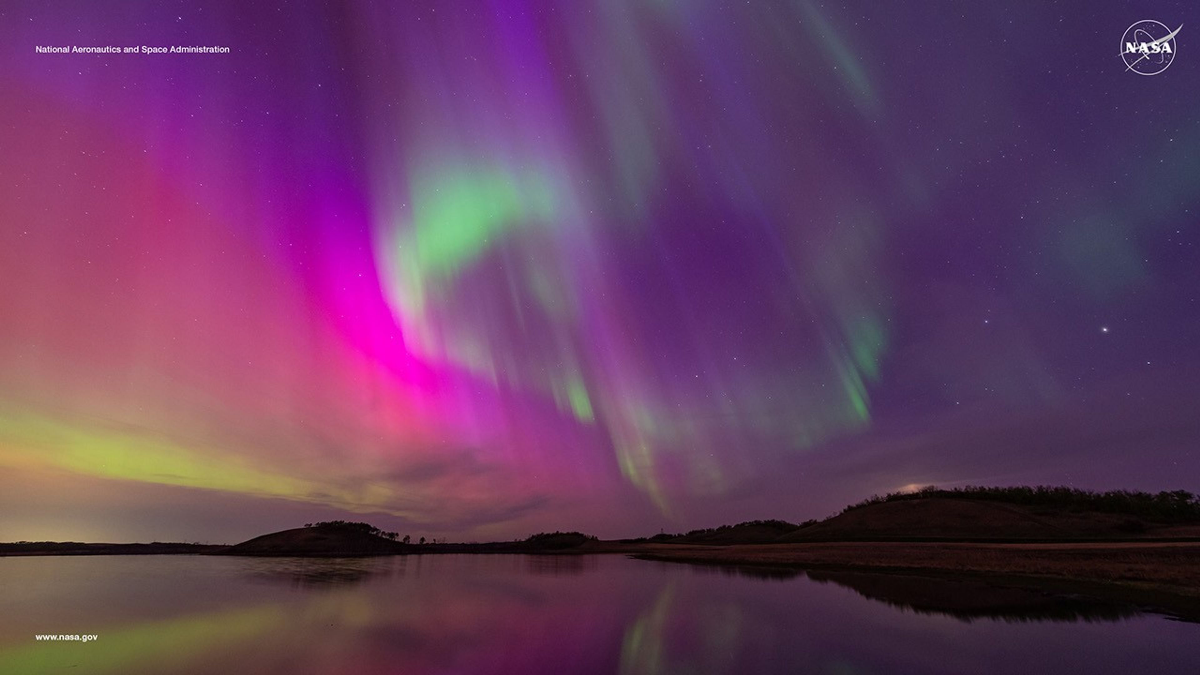 A vivid aurora borealis in shades of purple, green, and pink illuminates the night sky, reflected on a calm body of water with a silhouette of rolling hills in the distance. NASA logo and text 'National Aeronautics and Space Administration' are visible in the upper corners.