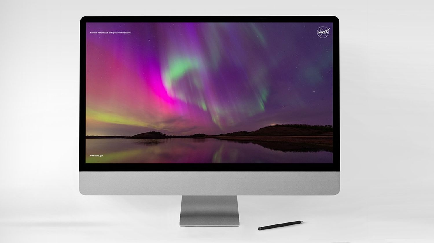 Desktop computer displaying a vibrant NASA-themed screen with a colorful aurora borealis reflecting over a calm body of water under a night sky.