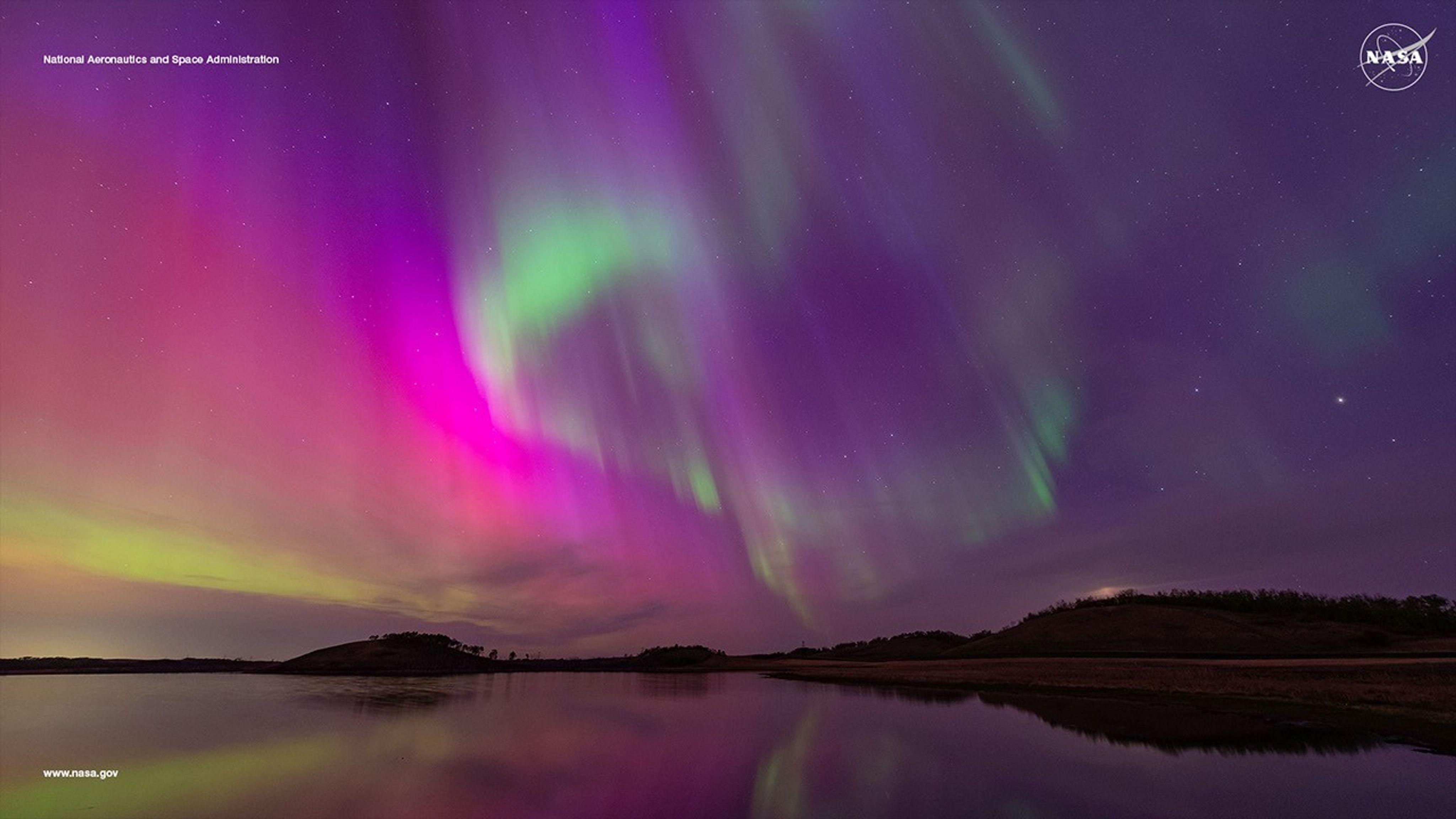 A vivid aurora borealis in shades of purple, green, and pink illuminates the night sky, reflected on a calm body of water with a silhouette of rolling hills in the distance. NASA logo and text &#039;National Aeronautics and Space Administration&#039; are visible in the upper corners.