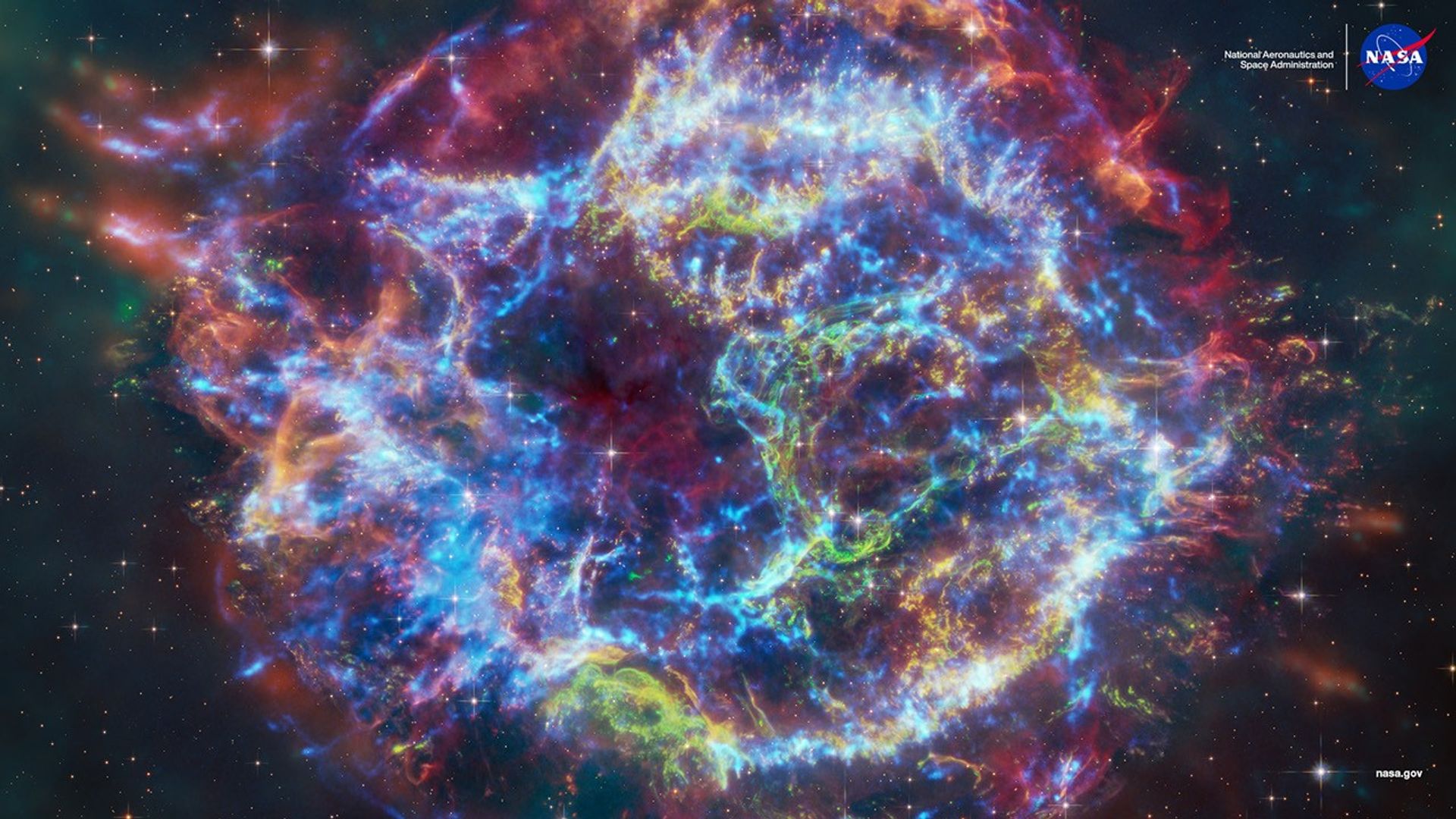 Colorful composite image of the supernova remnant Cassiopeia A, showing intricate, glowing filaments in blue, red, green, and yellow. Captured using NASA’s James Webb and Chandra telescopes, the image reveals expanding shockwaves and stellar debris from the exploded star, set against a star-filled background.