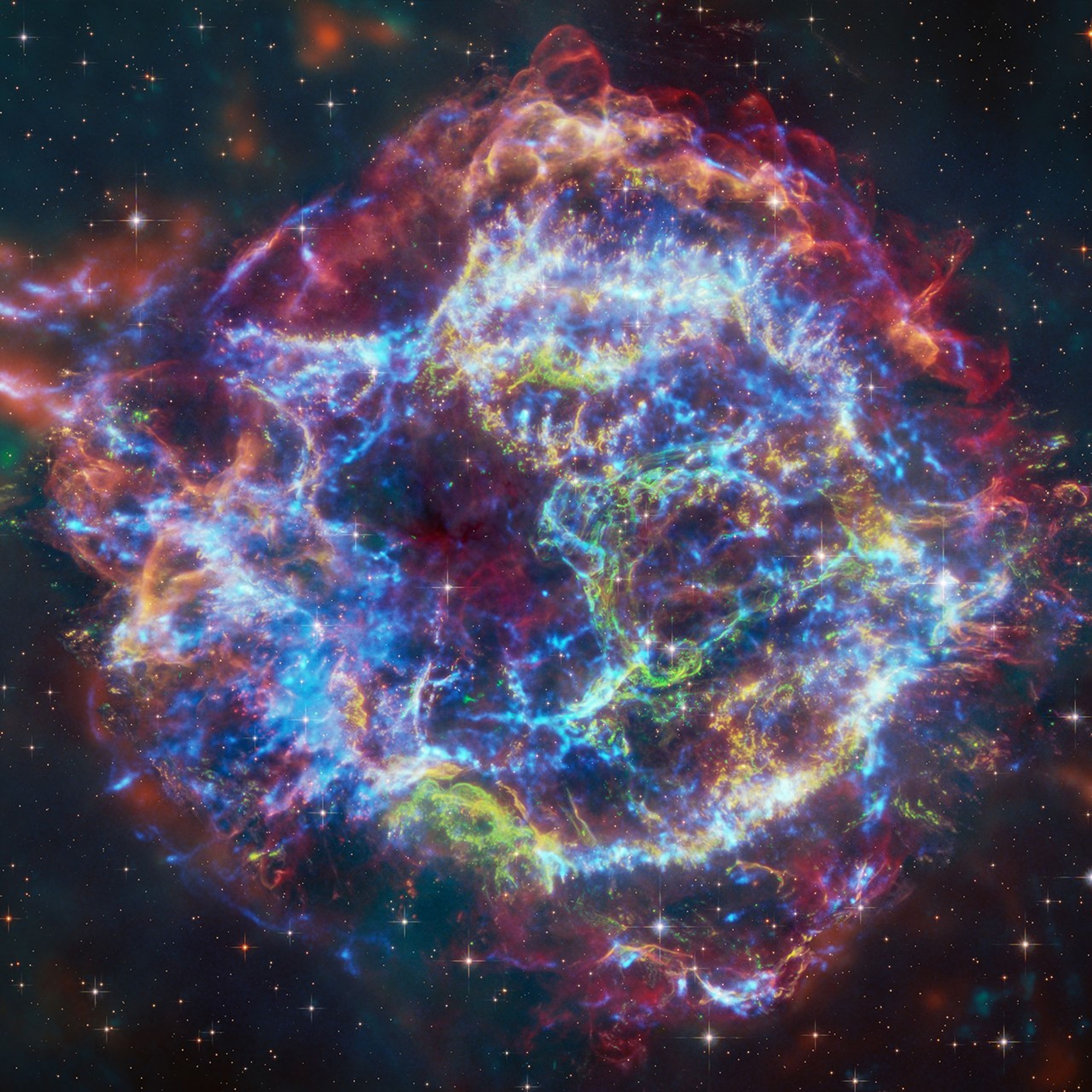 Colorful composite image of the supernova remnant Cassiopeia A, showing intricate, glowing filaments in blue, red, green, and yellow. Captured using NASA’s James Webb and Chandra telescopes, the image reveals expanding shockwaves and stellar debris from the exploded star, set against a star-filled background.